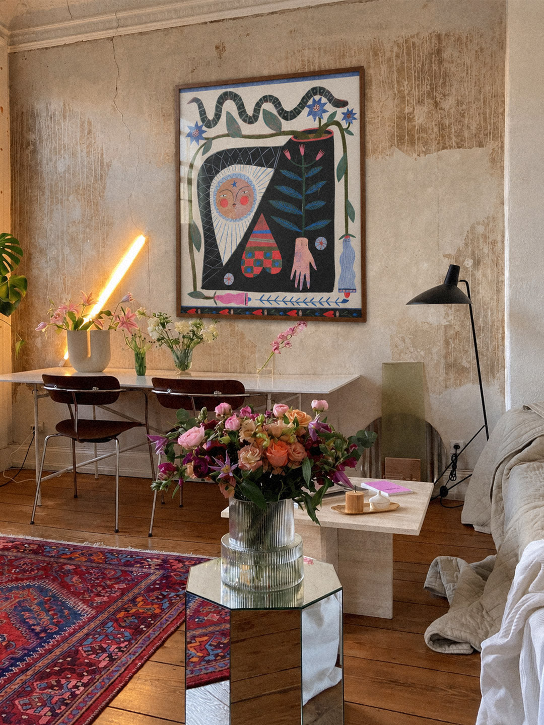 A framed abstract art print featuring stylized figures and organic shapes in black, blue, pink, and cream, hanging on a textured plaster wall in a rustic, modern living room with a wooden floor, red patterned rug, and floral arrangements. Poster produced by DAE