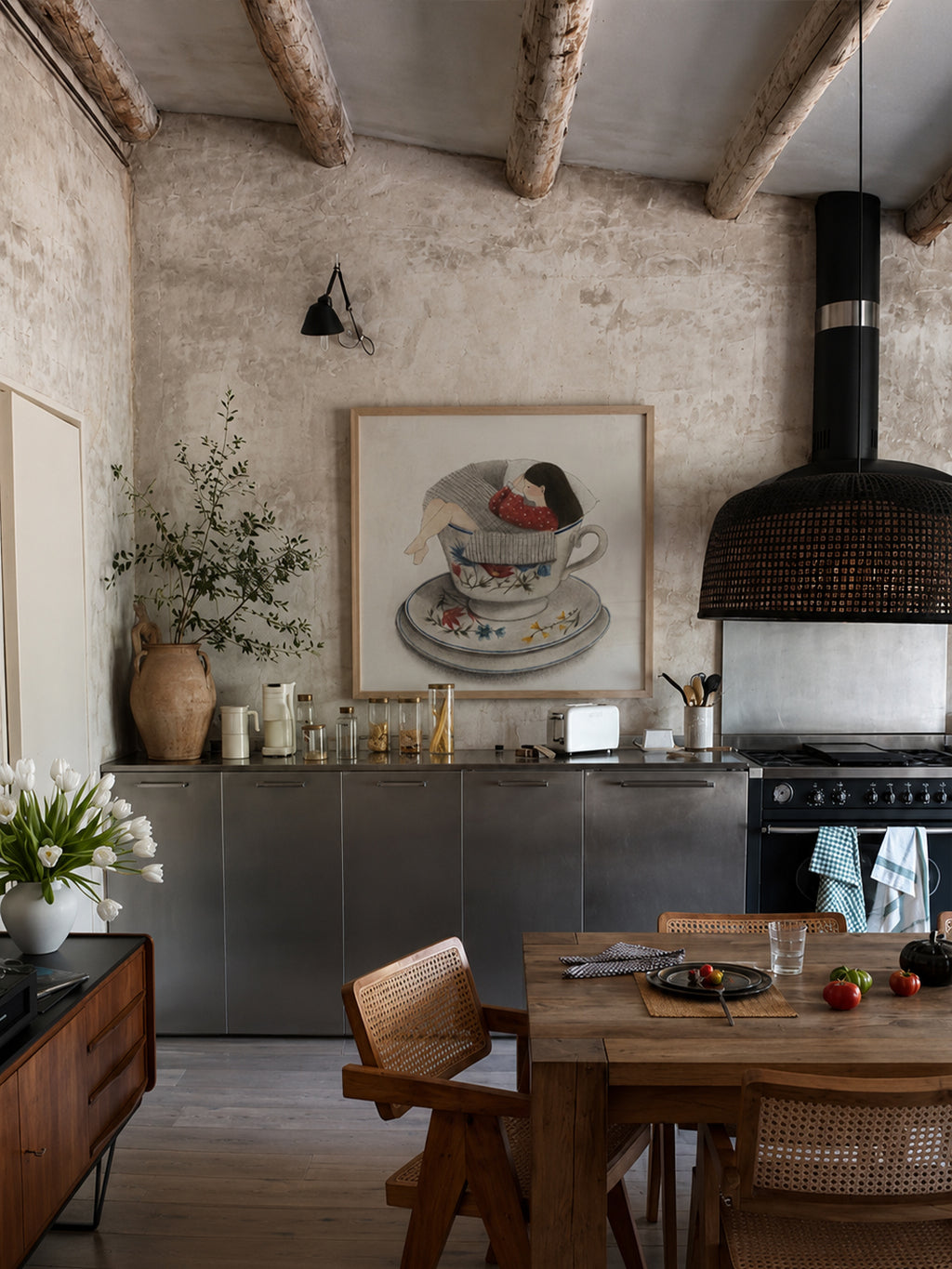 A rustic kitchen interior with a large framed art print by DAE depicting a teacup illustration hanging on a textured plaster wall. The room features exposed wooden ceiling beams, a dark wood dining table with woven chairs, a stainless steel range hood, and a rustic wooden sideboard.