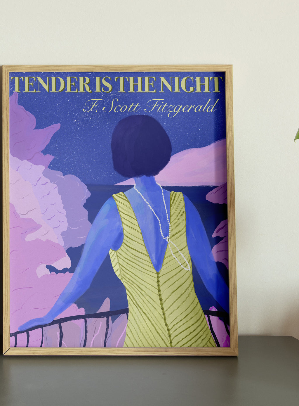A framed poster of a woman in a yellow dress viewed from behind, looking out over a railing with a night sky and city lights in the background. The title 'Tender is the Night' by F.