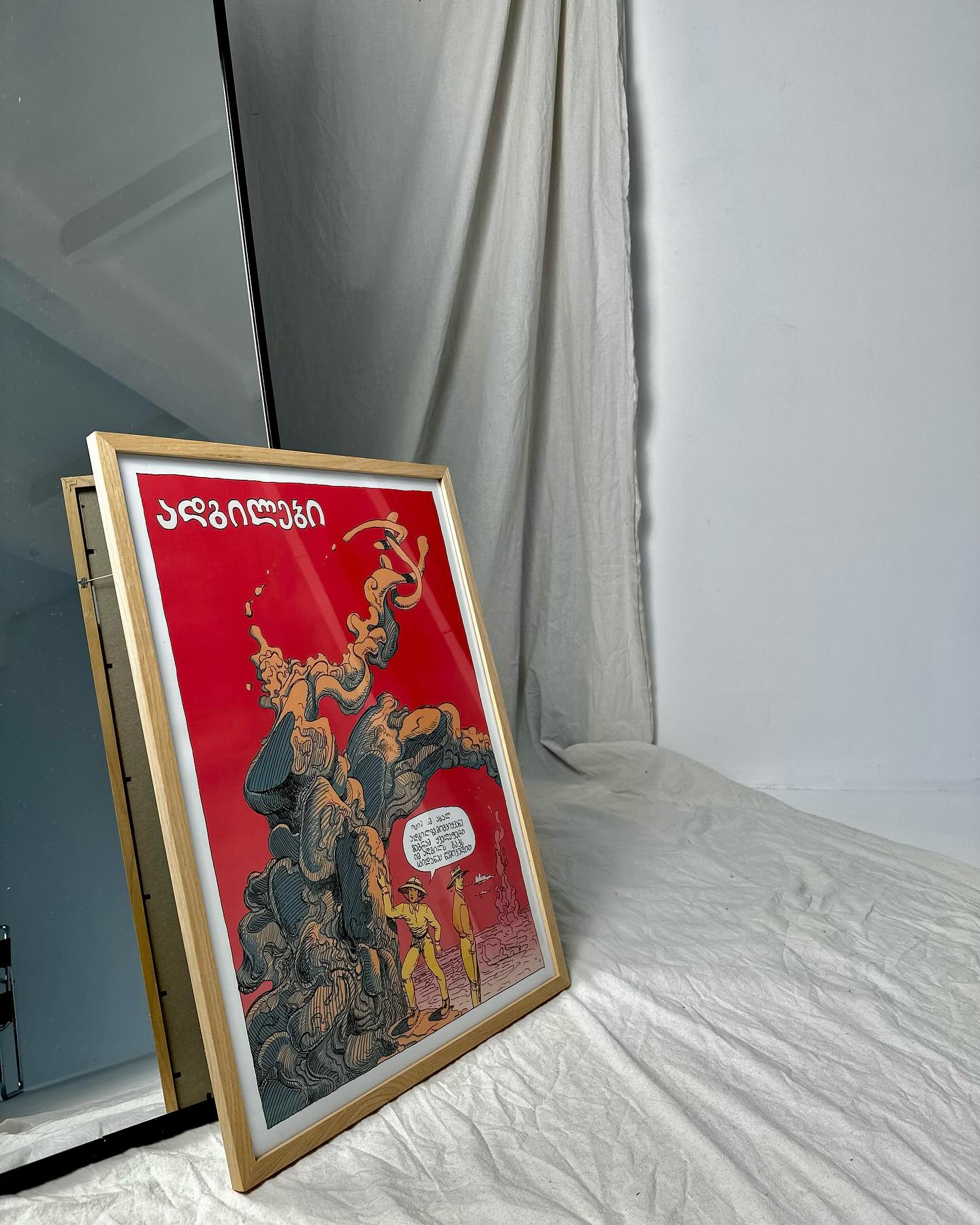 poster with georgian words standing on the floor, next to it is mirror and plain, white, studio like background