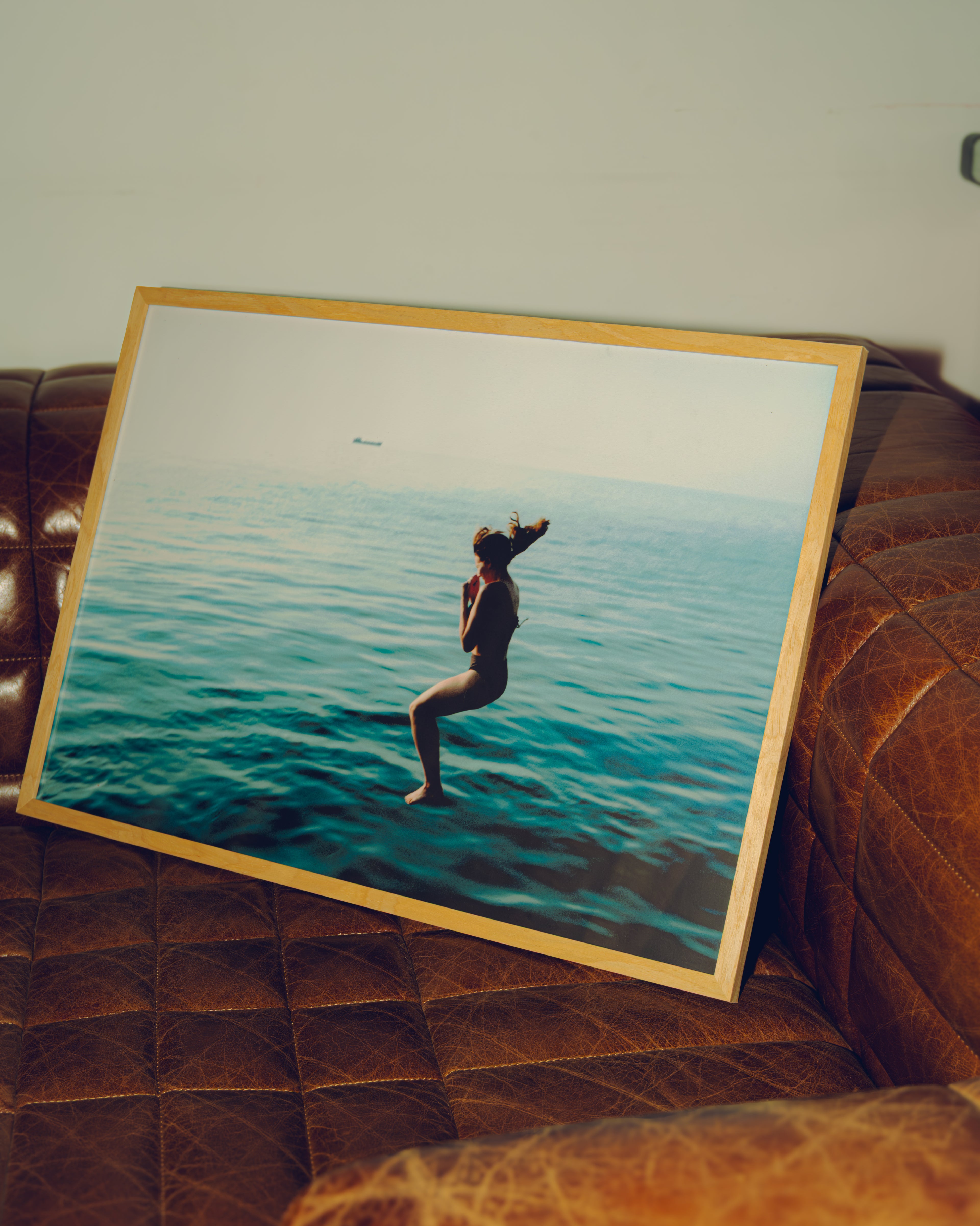 Framed artwork of a person jumping into the water leaning against a brown leather couch.