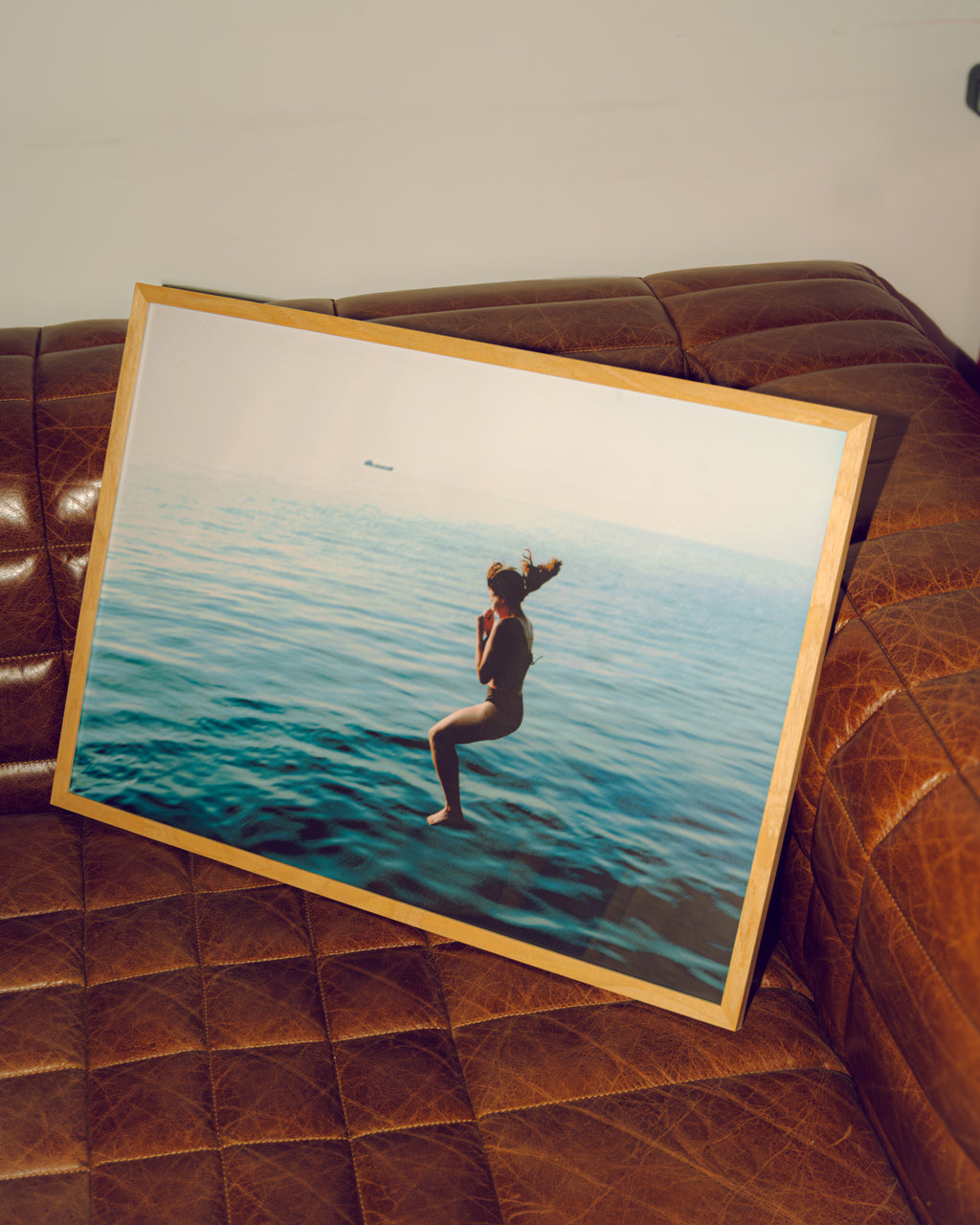 Framed artwork of a person jumping into the water with a sailboat in the background, placed on a brown leather couch.