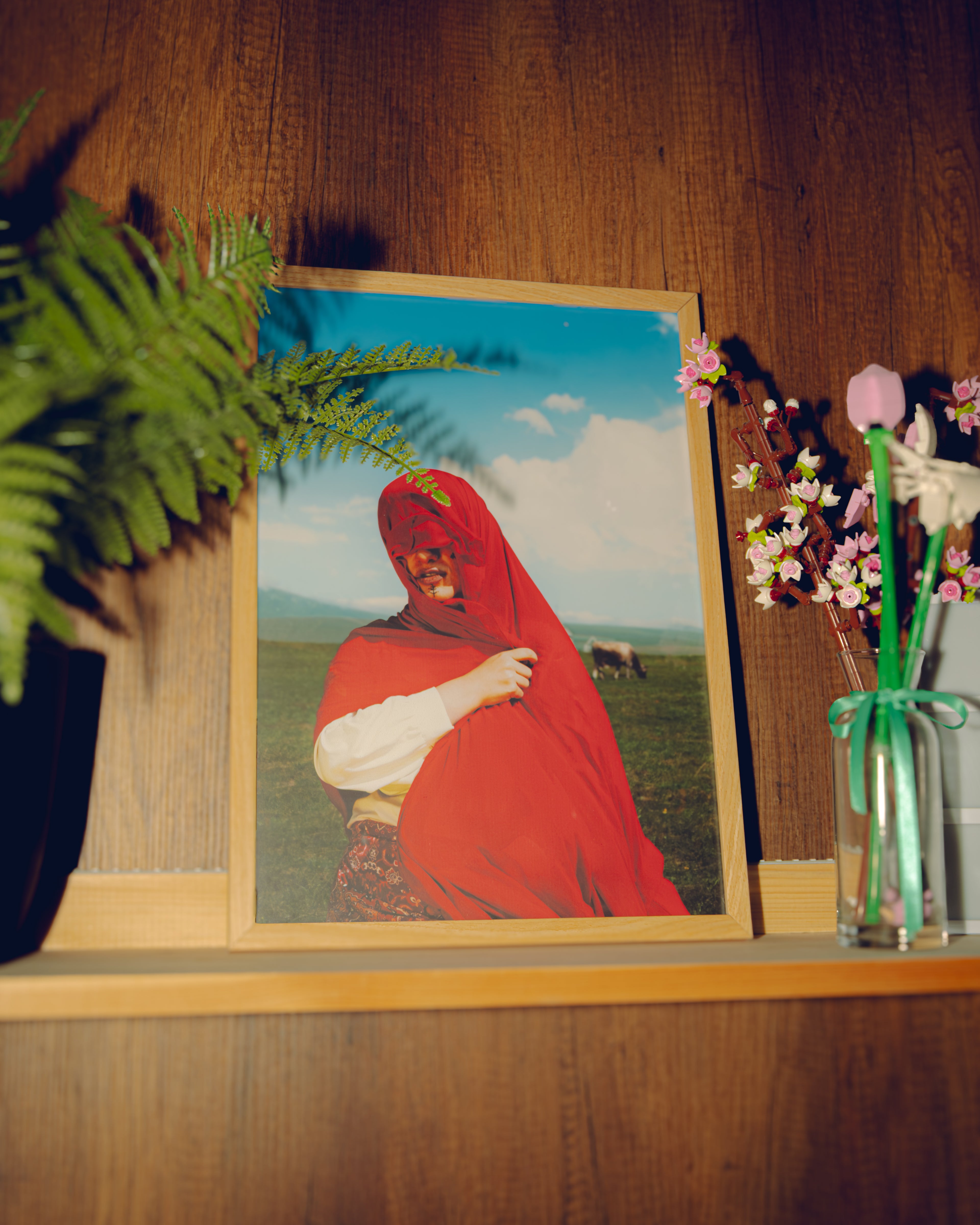 Framed picture of a person in a red garment on a wooden shelf with plants and flowers.