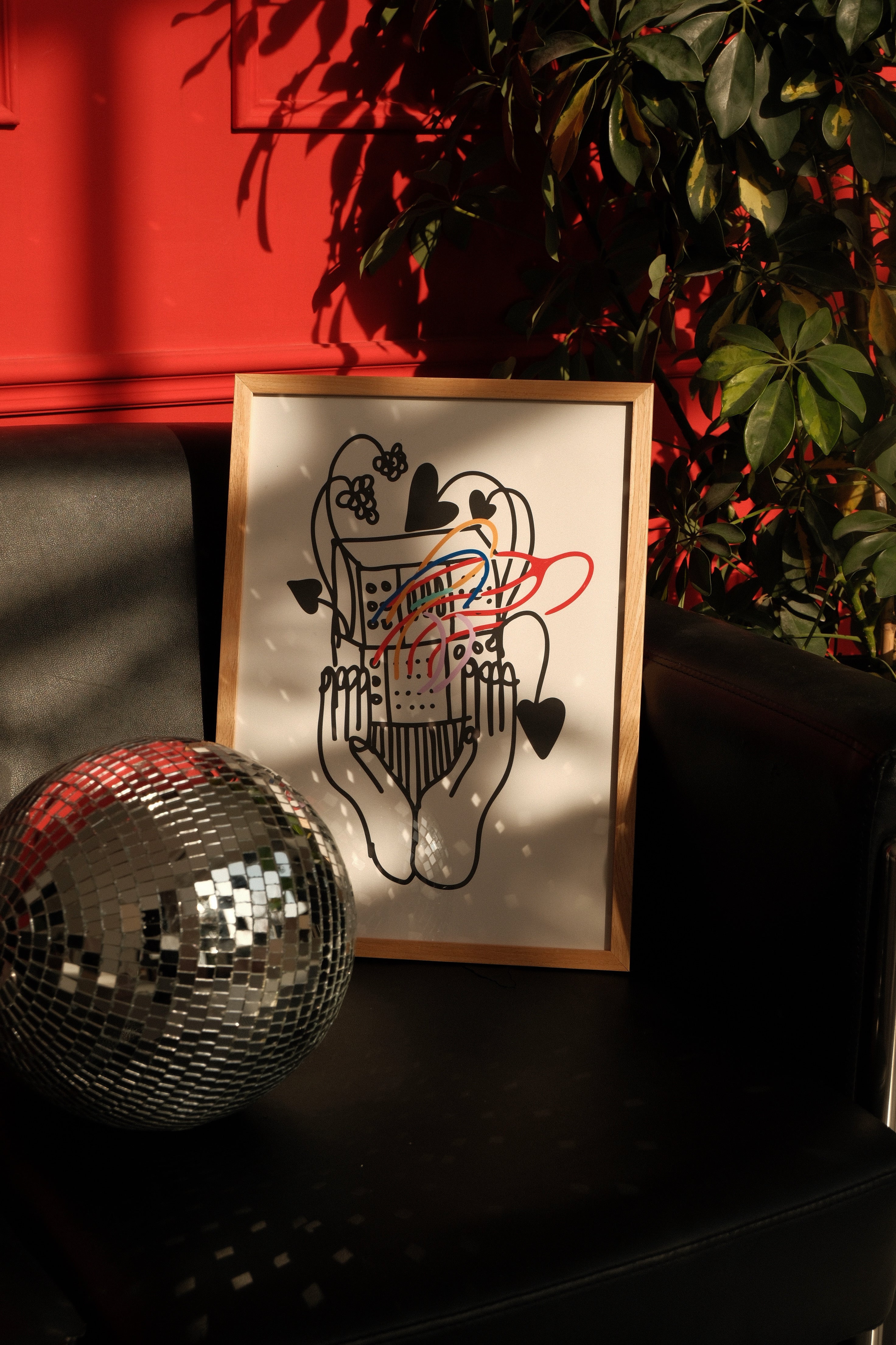 Framed artwork with abstract design on a dark surface next to a disco ball, with a red wall and plant in the background.