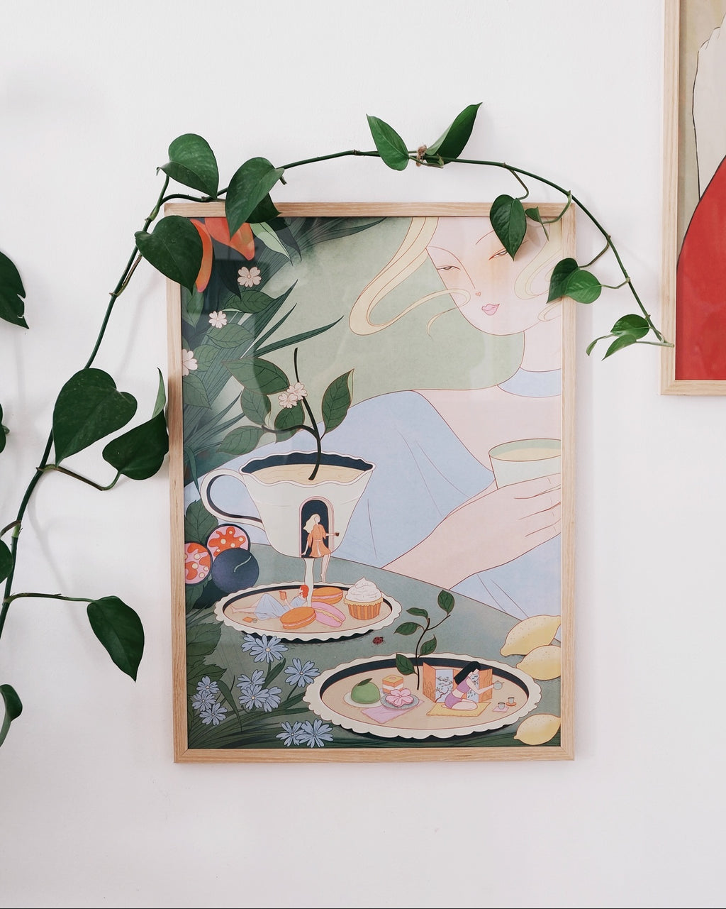 Poster “Afternoon Tea” by Leah Han, displayed in wooden frame on wall with plants
