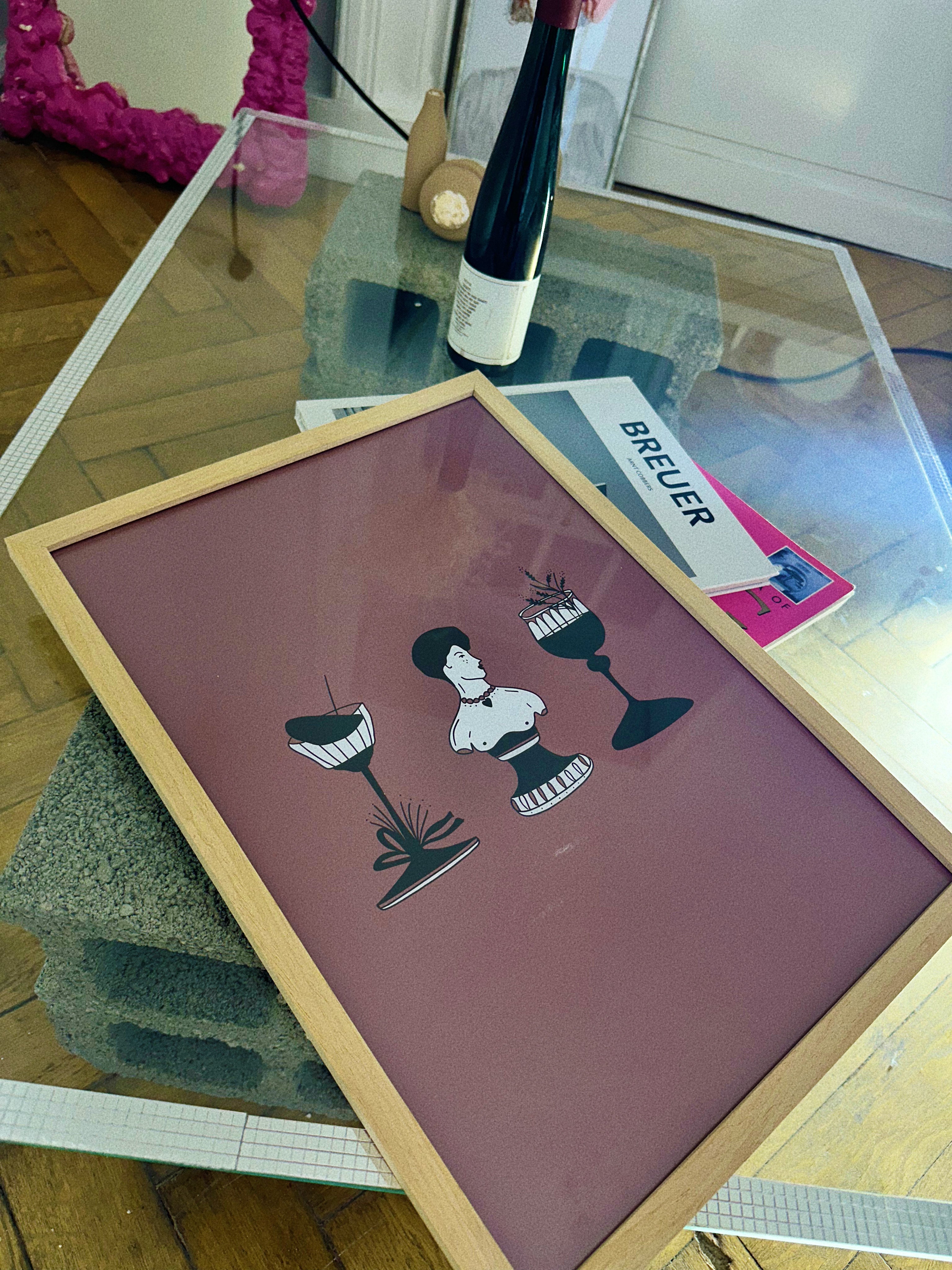 A framed poster featuring a stylized illustration of a person and a cocktail glass, displayed on a table with various objects.