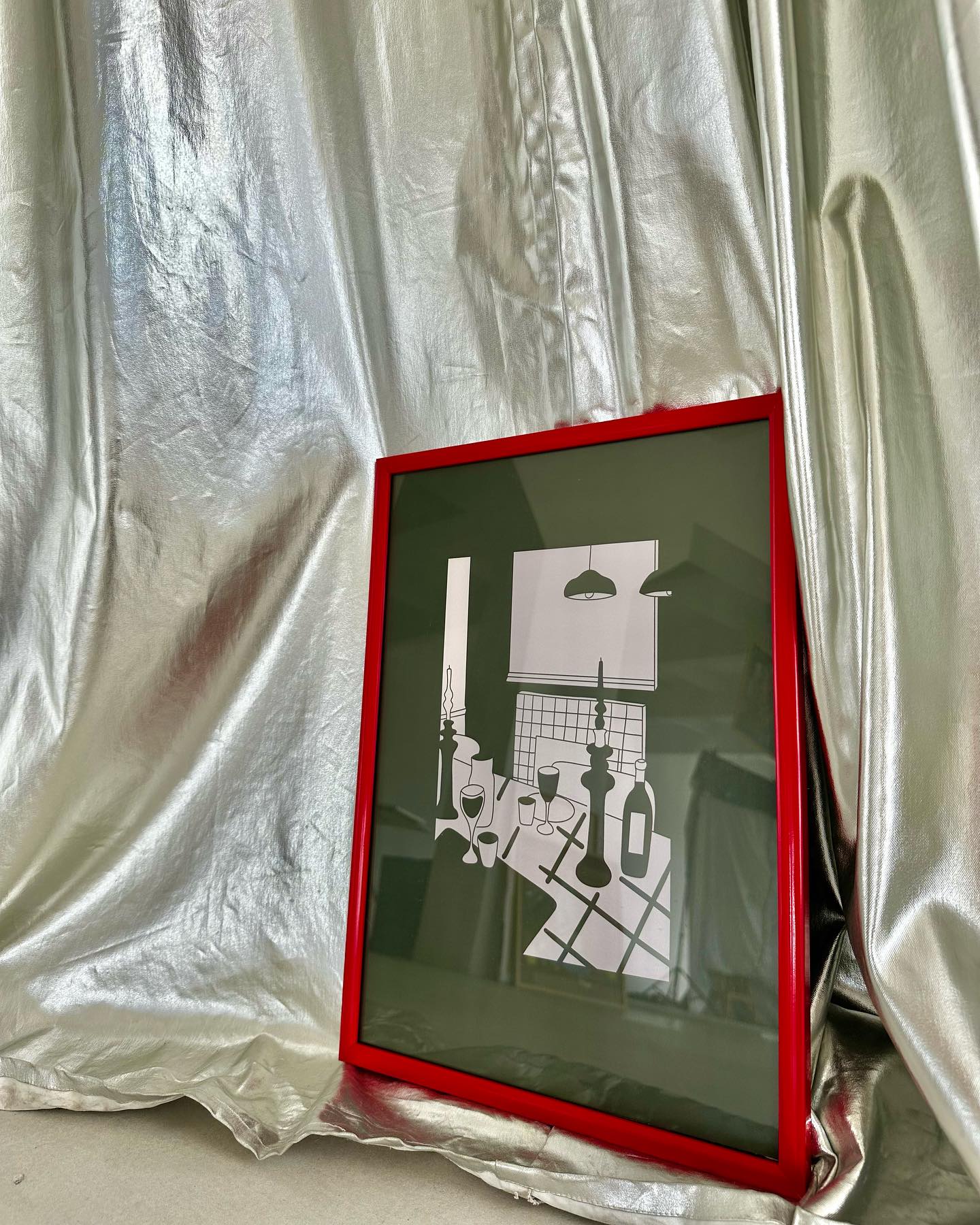 A poster depicting a stylized dining room setting with a red frame, displayed against a silver fabric background.