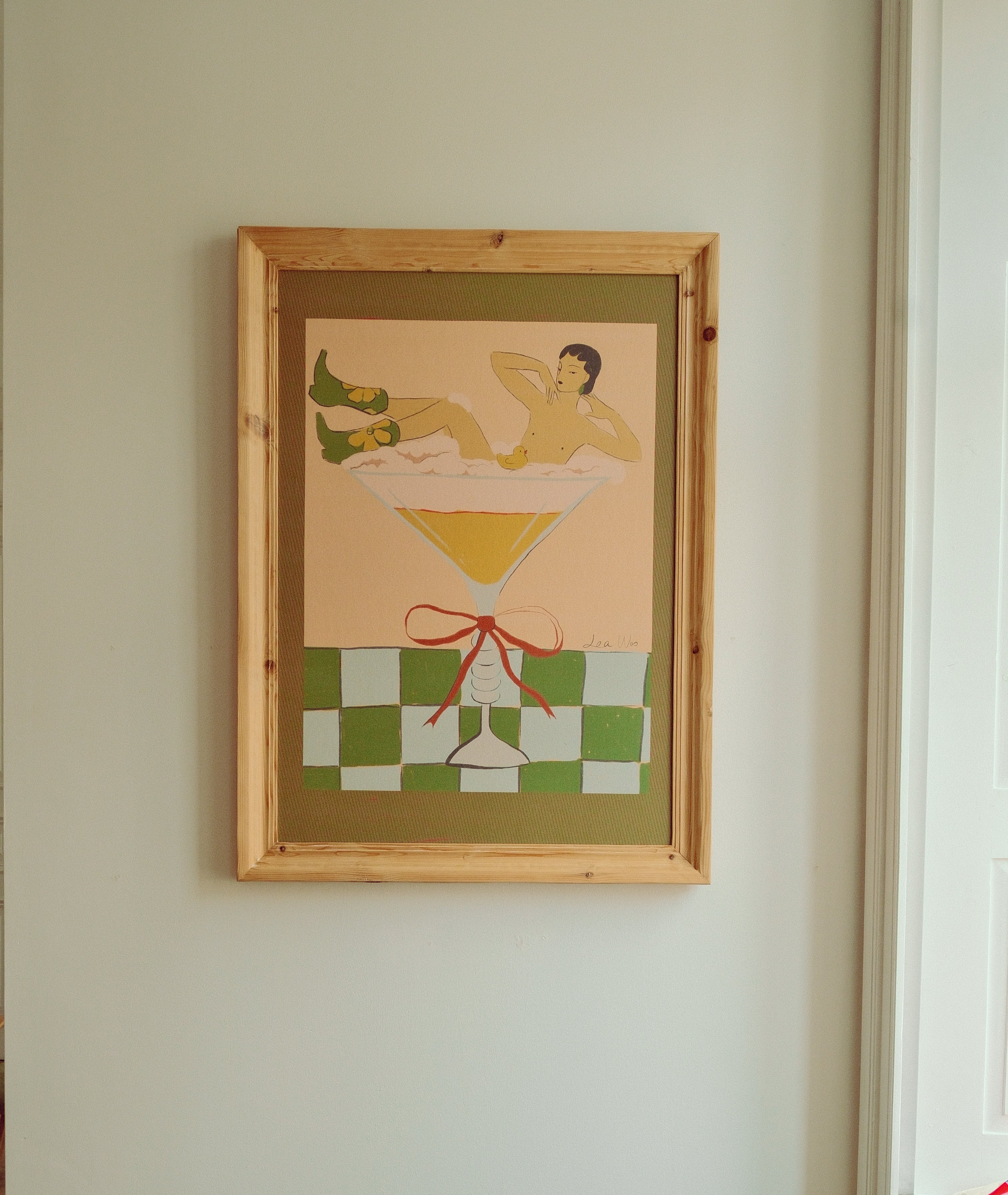 A framed art print featuring a retro illustration of a woman lounging in a martini glass, hanging on a light-colored wall.