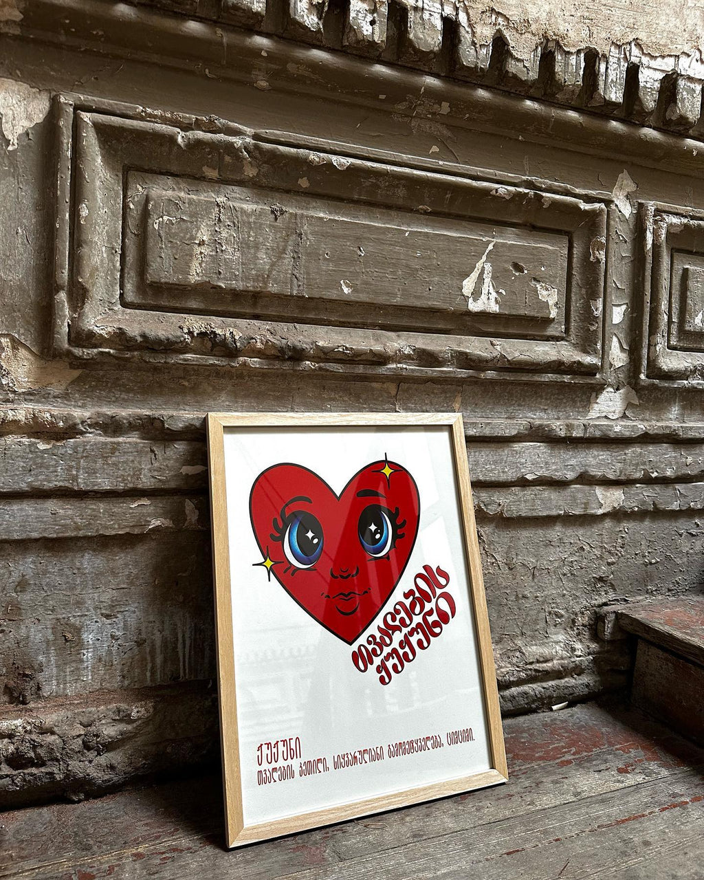 A framed poster by DAE with a red heart and a face illustration inside it, placed against an old, textured wall.