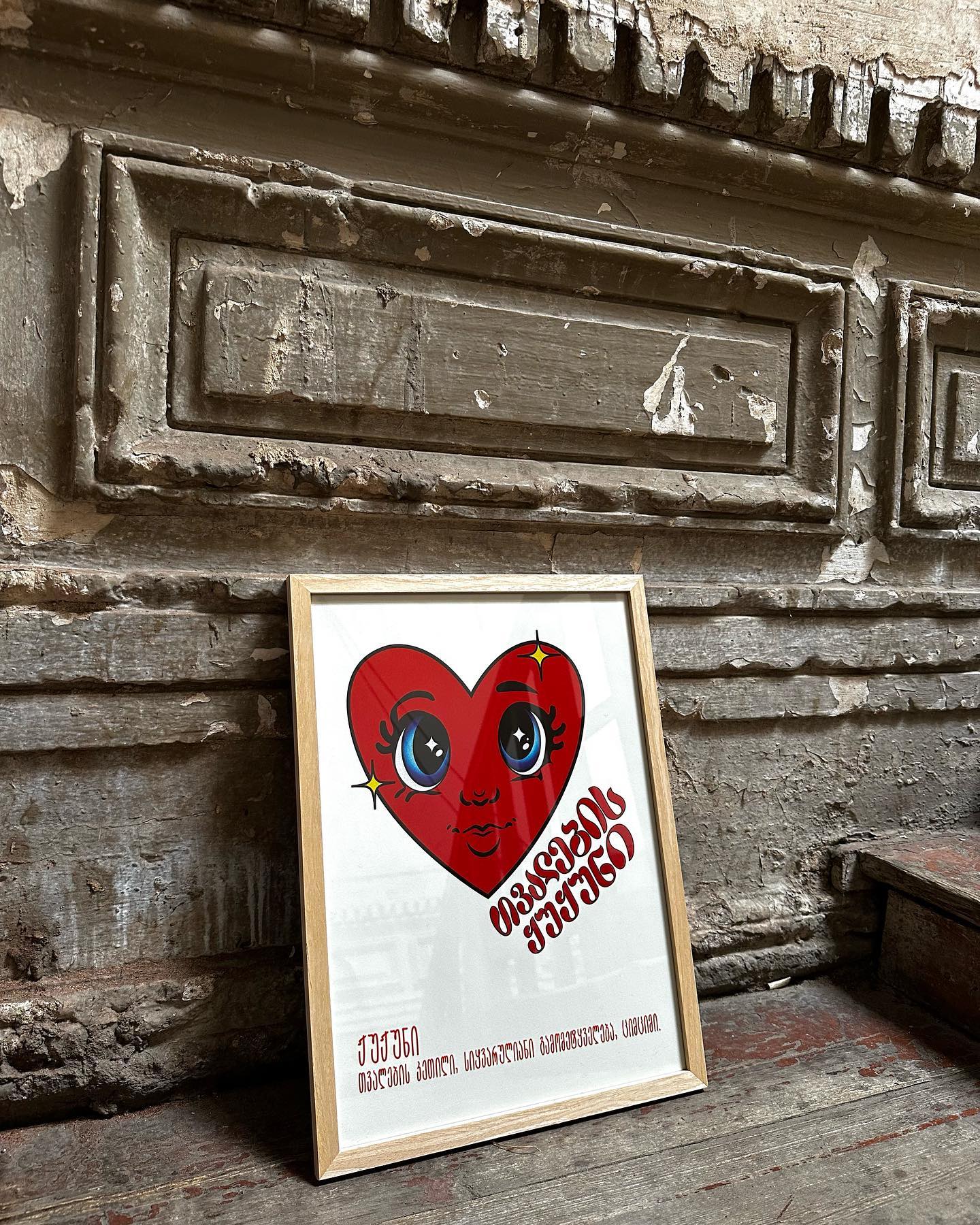 A framed poster by DAE with a red heart and a face illustration inside it, placed against an old, textured wall.