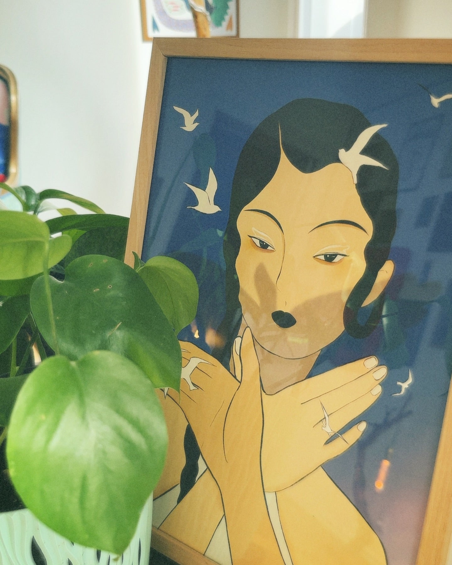 A poster featuring a stylized illustration of a woman with birds around her, set against a blue background. The poster is displayed next to a green potted plant.