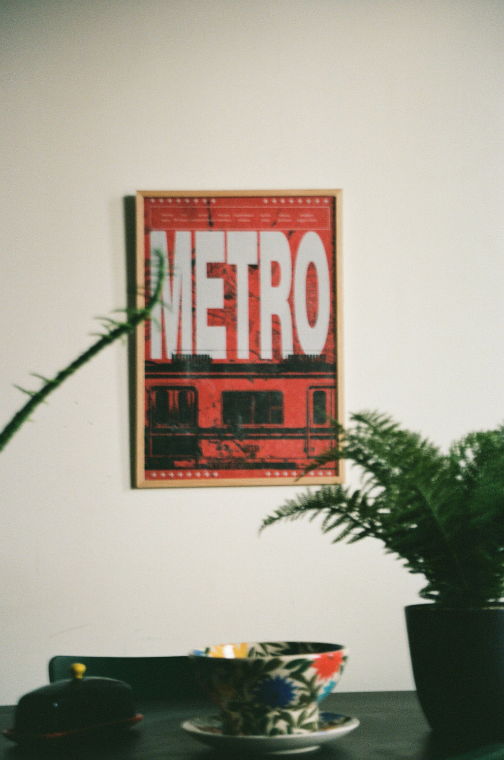 Framed poster with 'METRO' text on a wall with greenery. Tbilisi Metro poster with list of metro stations and city map background by Lasha Lezhava x DAE