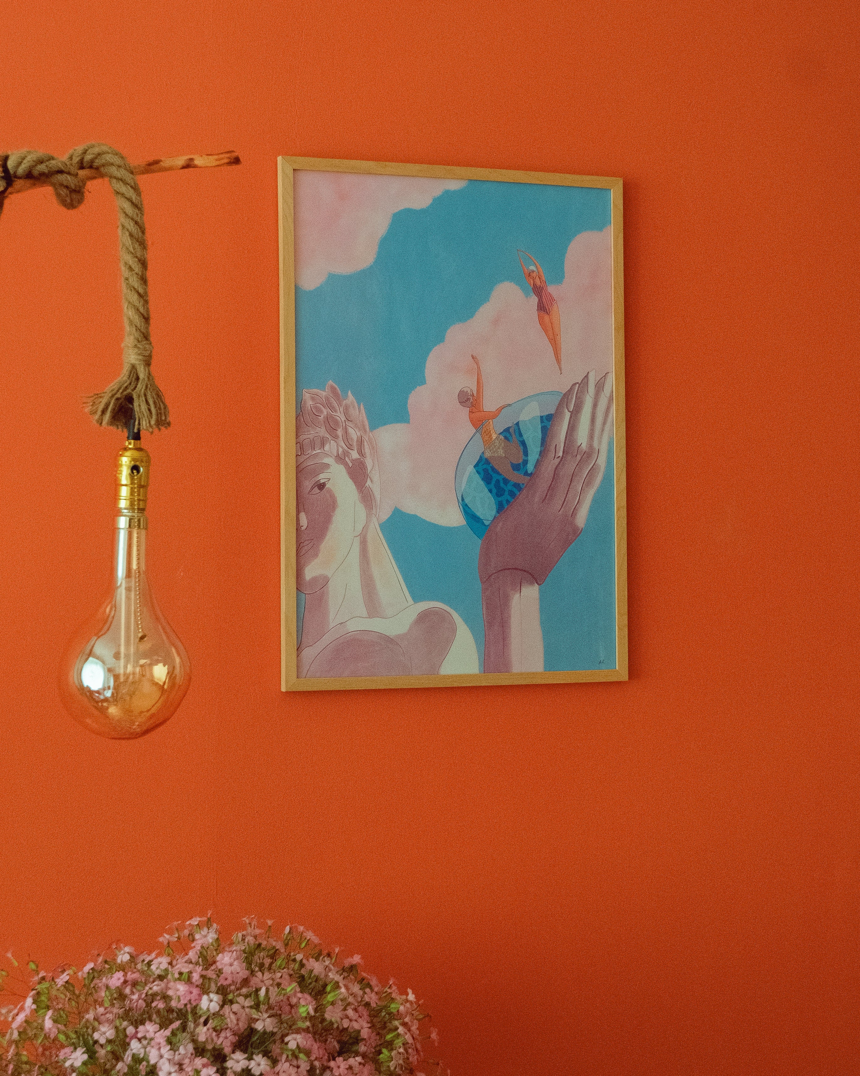 Framed artwork on an orange wall with a hanging light bulb and plant.