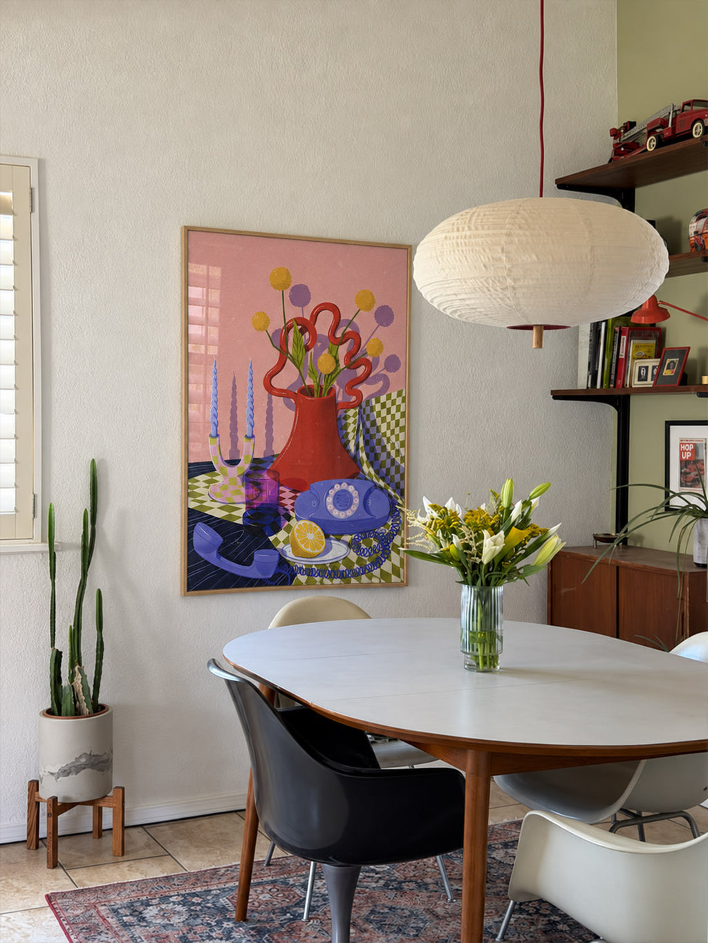 A colorful framed still life painting hangs on a beige wall above a mid-century modern dining table with chairs and a hanging pendant light in a styled dining room.