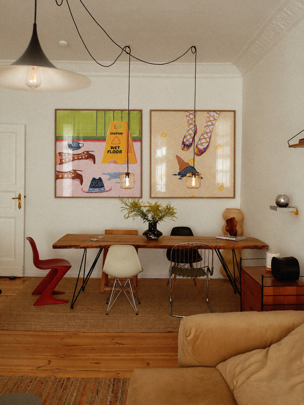 A styled interior room with two framed colorful art prints on a white wall above a wooden dining table, chairs, pendant lights, and a beige sofa in the foreground.