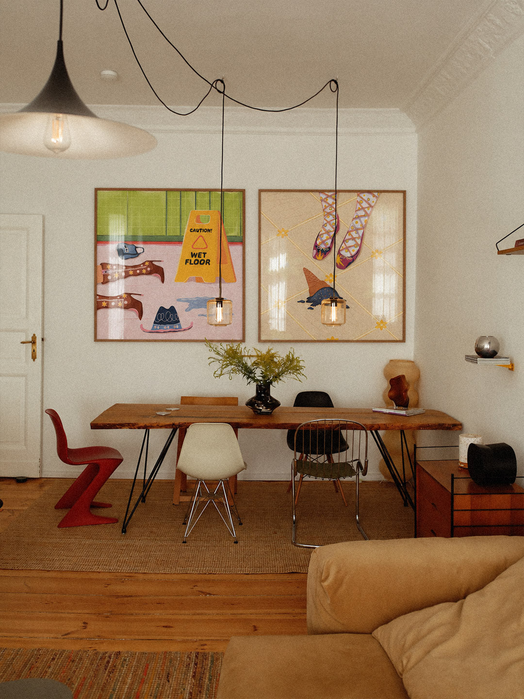 A styled interior room with two framed colorful art prints on a white wall above a wooden dining table, chairs, pendant lights, and a beige sofa in the foreground.