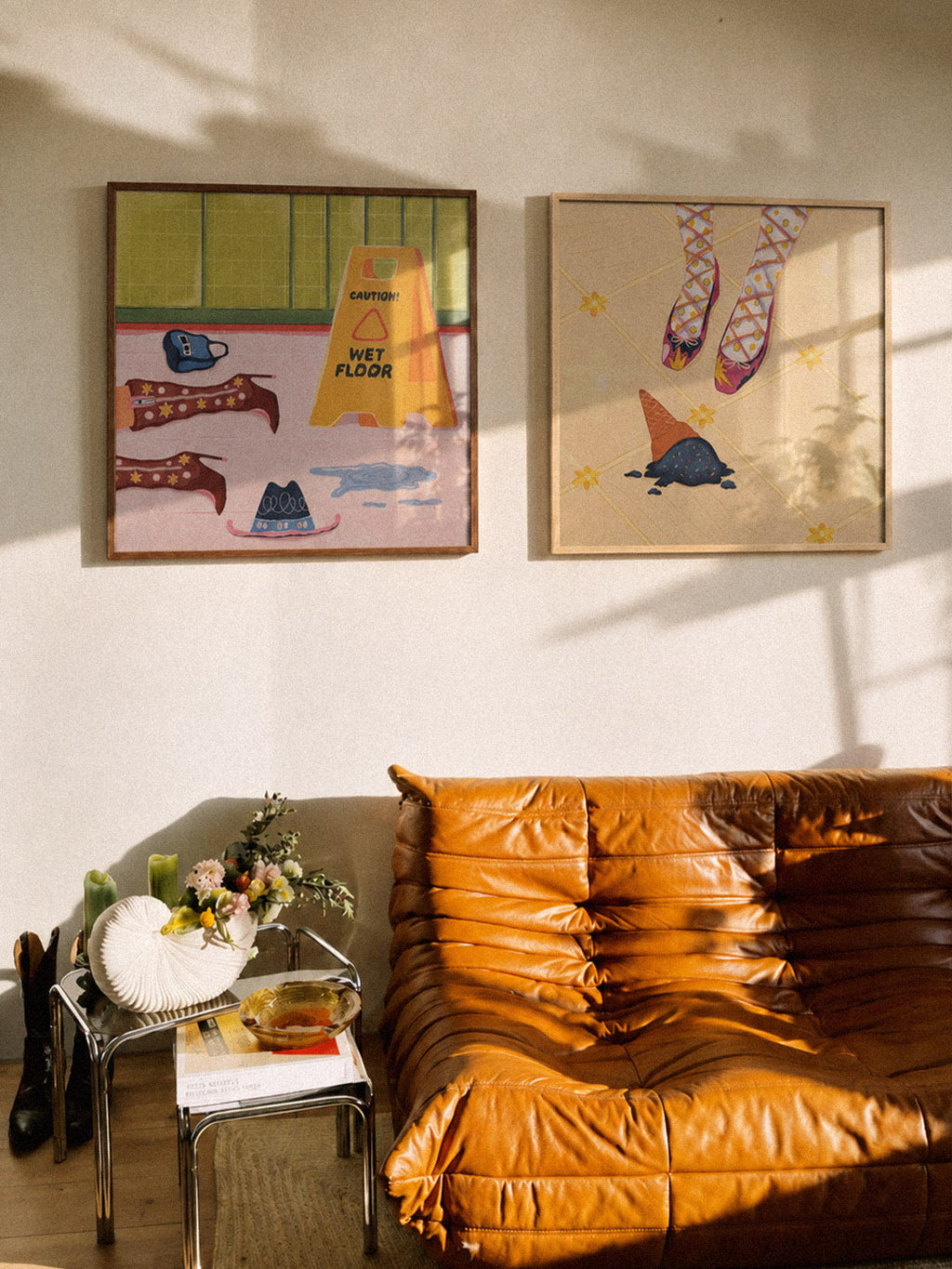 Two framed DAE's art prints hanging on a beige wall above a brown leather sofa in a sunlit room. The left print has pastel pink, green, and yellow colors, and the right print shows purple shoes and an ice cream.