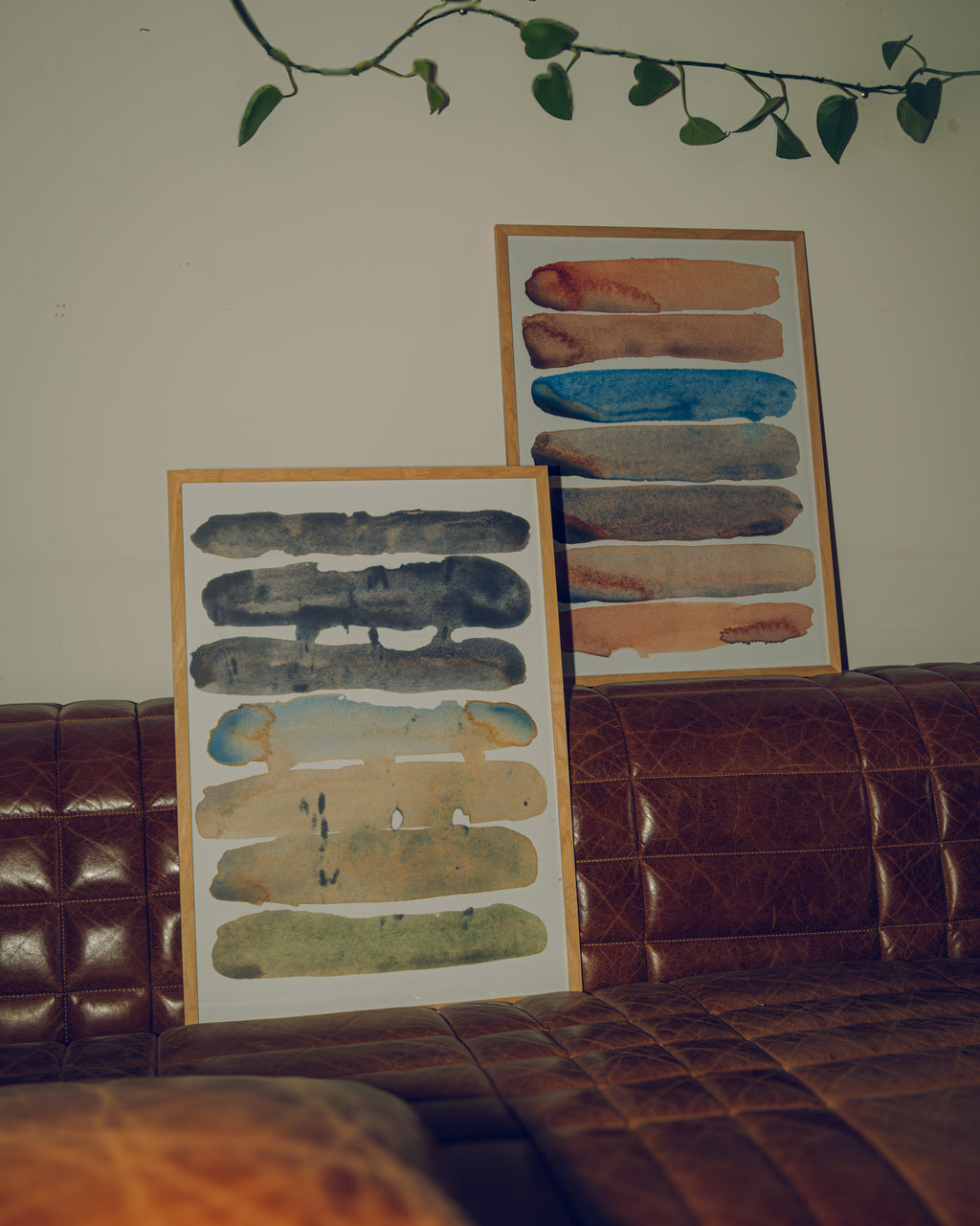 Two framed art pieces on a brown leather couch with a decorative plant above.