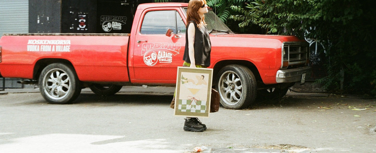 Woman standing next to a red truck with poster in her hand by DAE, with a store in the background