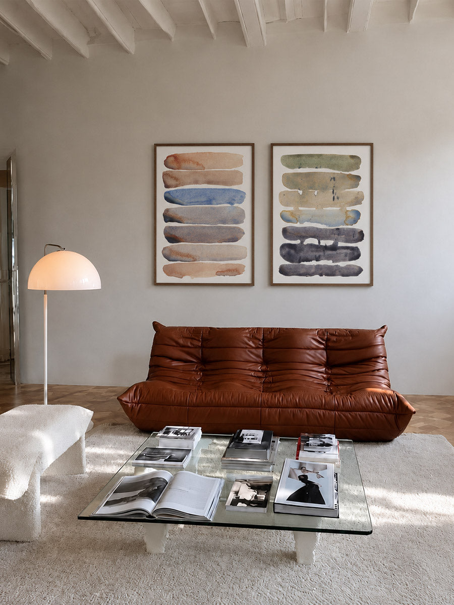 A modern living room featuring a rust-brown leather chaise lounge sofa, a glass coffee table with magazines, a white ottoman, and two framed abstract art prints by DAE on a light gray wall.