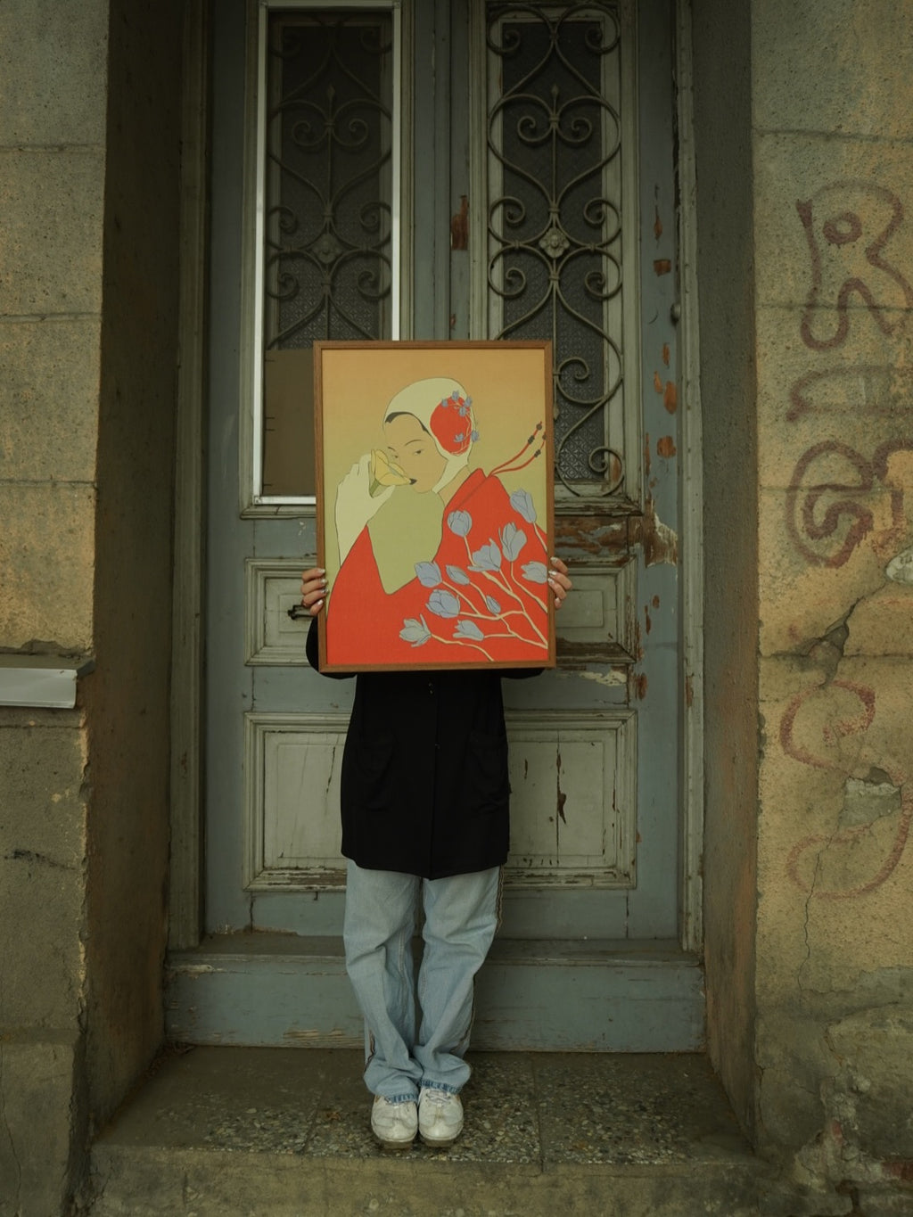 Person holding a framed art poster illustrated by Lea Woo, standing in front of an old wooden door. The poster features a woman with flowers and red tones.
