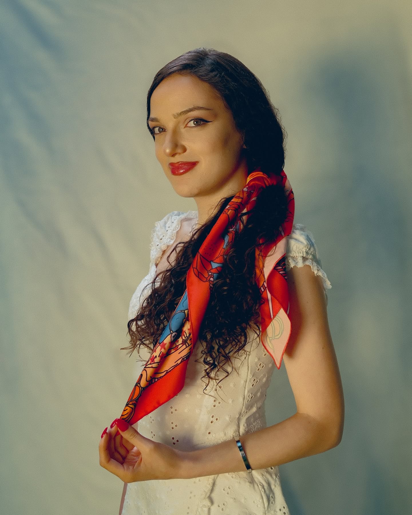 A woman with long dark curly hair wearing a white lace-trimmed dress and a vibrant red patterned silk scarf, posing against a soft, textured gray studio backdrop.