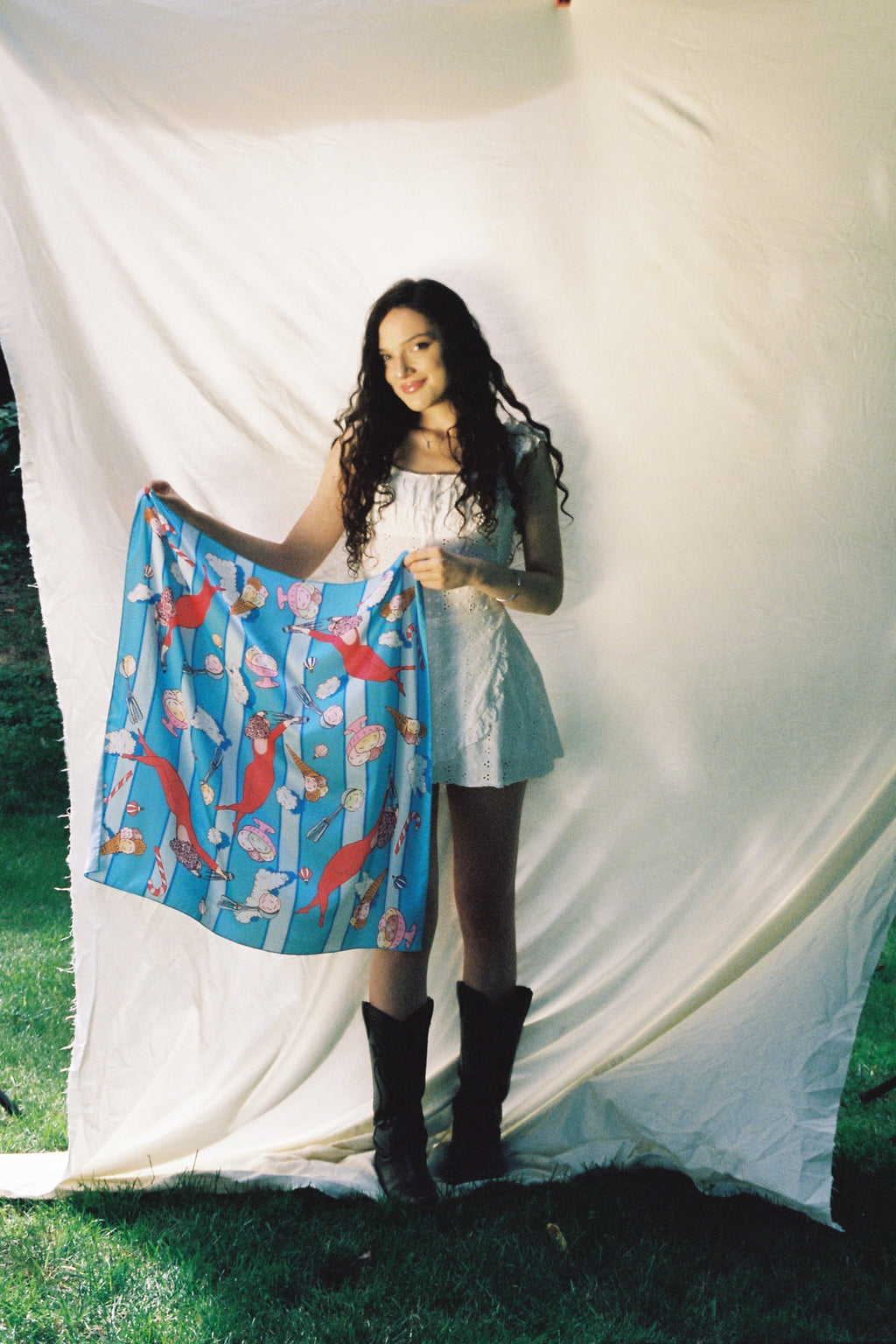 A woman lies on a large white sheet outdoors, holding up a blue patterned scarf item with red, white, and yellow abstract motifs. She is wearing black knee-high socks and a white top, with grass visible around the sheet.