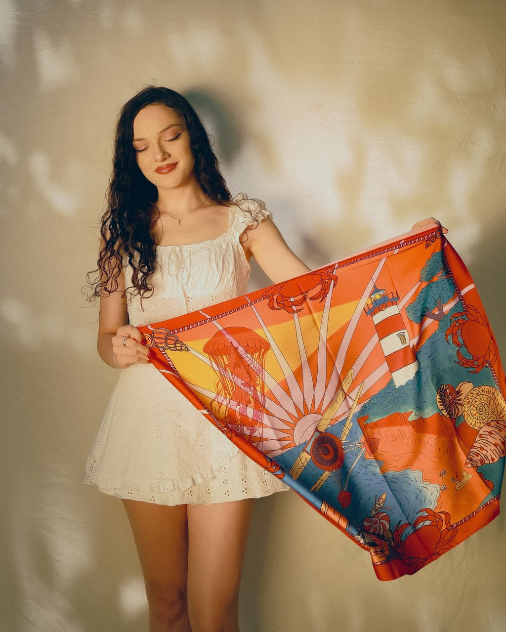 A woman in a white dress holds up a vibrant silk scarf featuring a colorful sun, lighthouse, and coastal landscape print in orange, yellow, blue, and red tones against a textured beige wall.