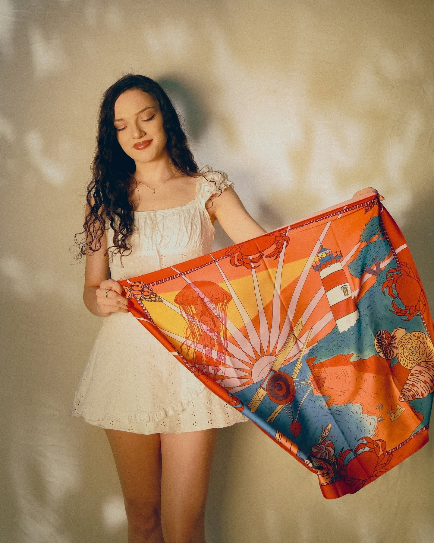 A woman in a white dress holds up a vibrant silk scarf featuring a colorful sun, lighthouse, and coastal landscape print in orange, yellow, blue, and red tones against a textured beige wall.