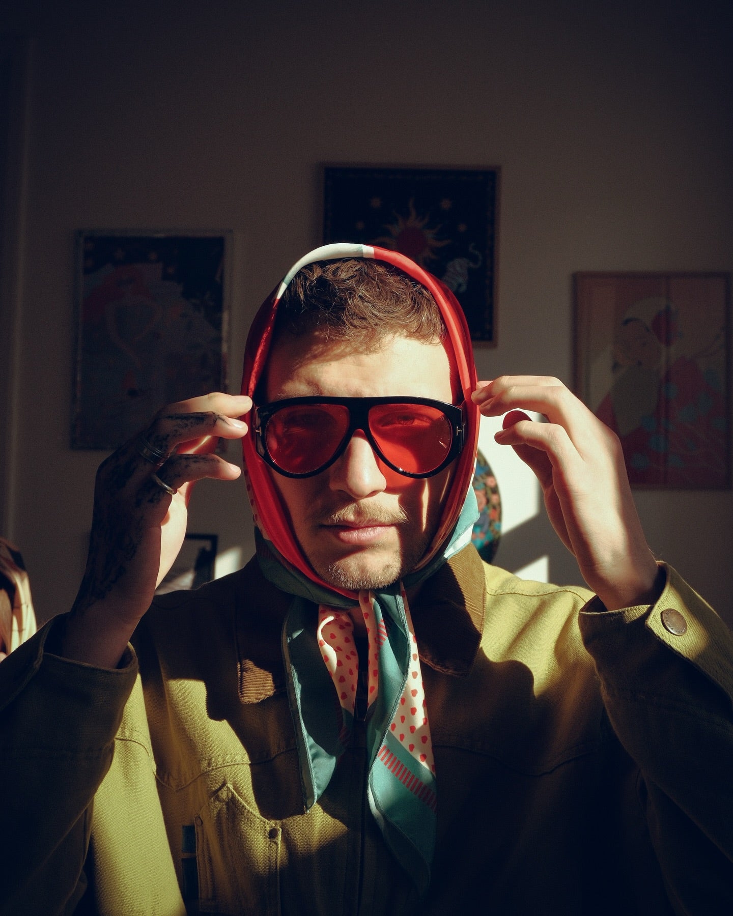 Person wearing a red hood and sunglasses in a room with artwork on the walls.
