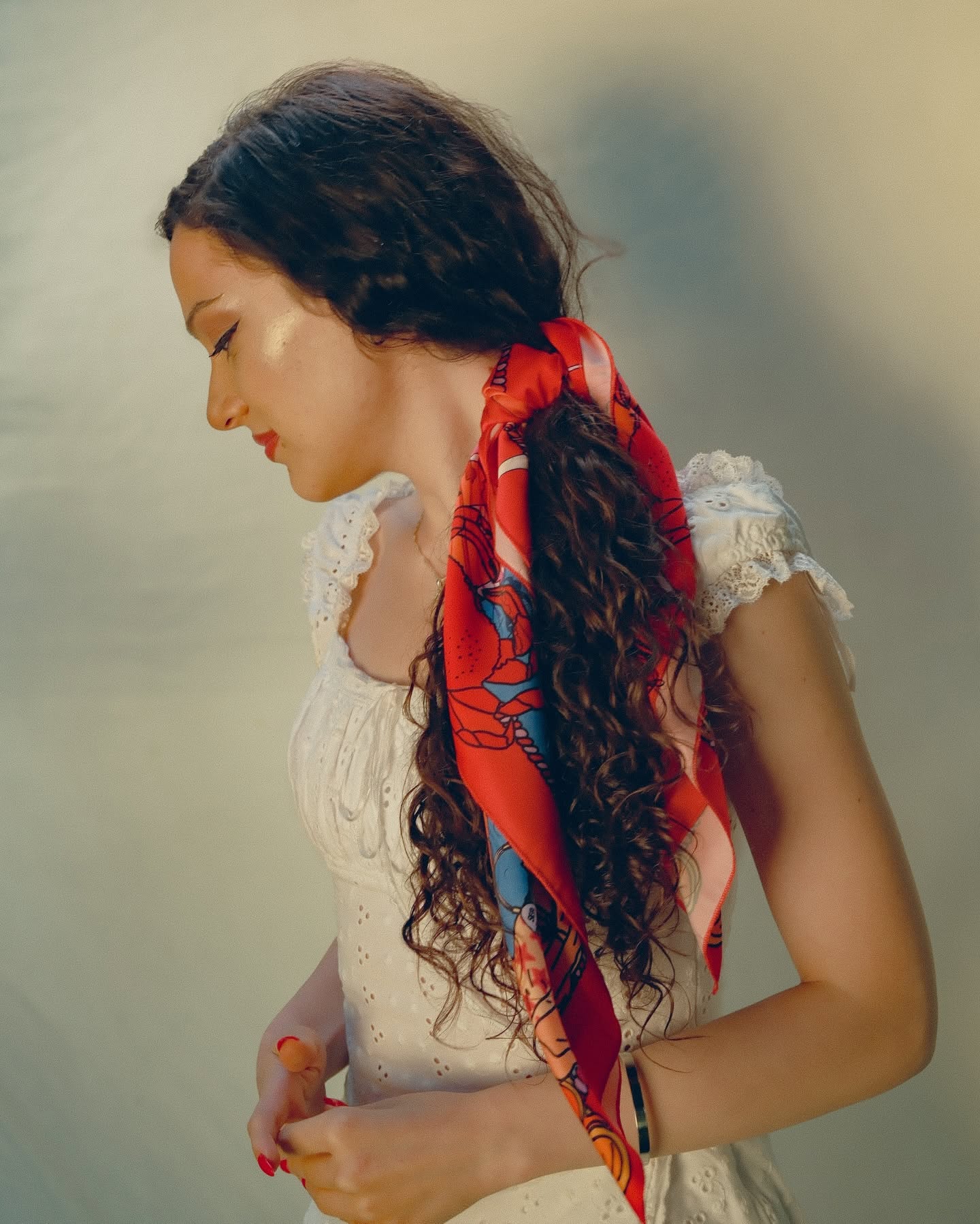 A woman with long, curly dark brown hair is shown in profile, wearing a white lace-trimmed dress and a vibrant red silk scarf with blue and orange patterns tied around her hair. She is posed against a soft, warm-toned, neutral background.