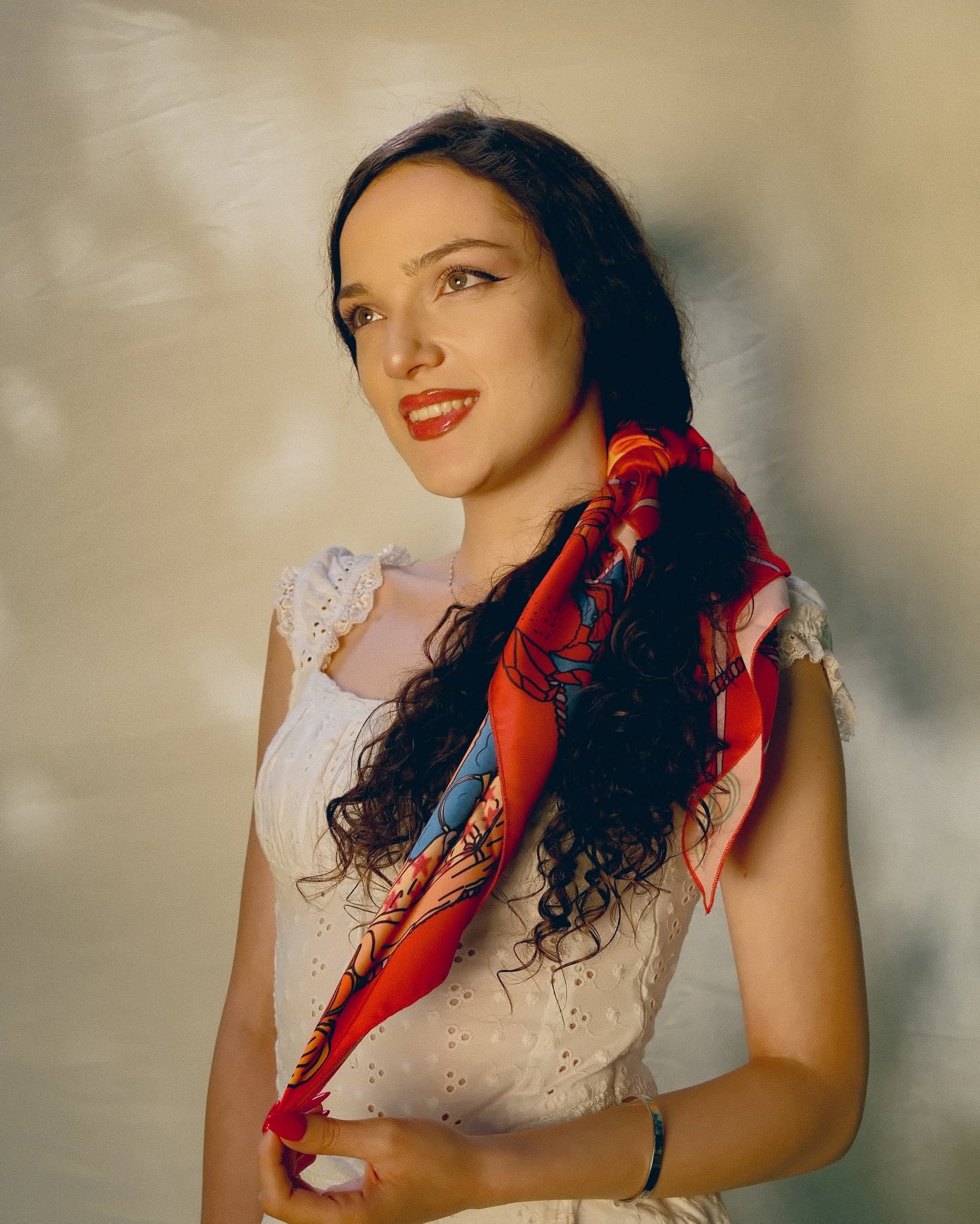 A woman with long dark curly hair wearing a white lace-trimmed top and a red patterned silk scarf, posed against a warm beige textured background.