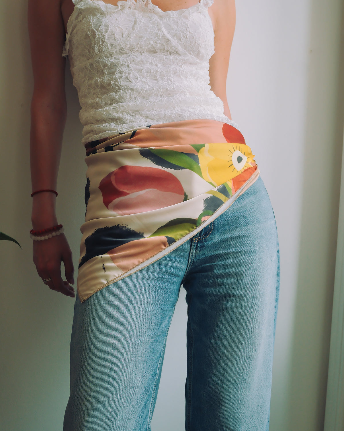 A person wearing a floral satin wrap scarf tied around the waist over a white lace camisole top and blue jeans.