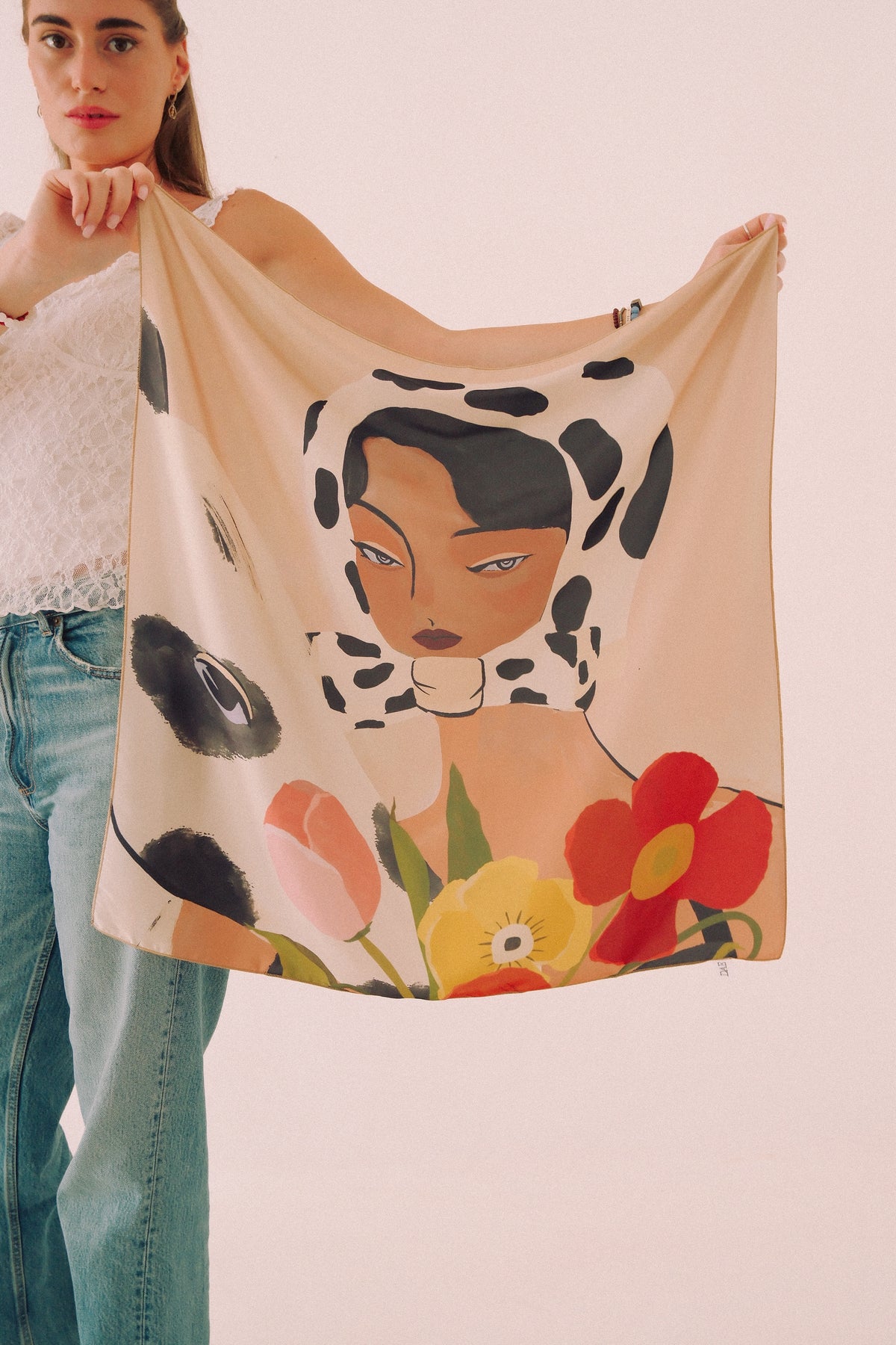 A woman holds up a square scarf featuring a stylized illustration of a woman in a black-and-white cow-print headscarf with colorful flowers at the bottom, against a plain light background.
