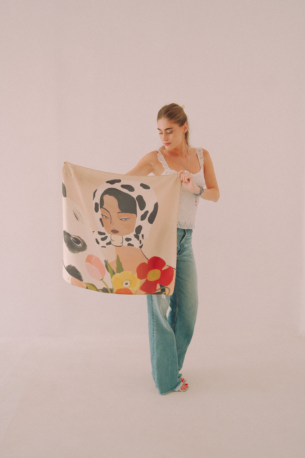 A woman holds up a large square scarf featuring a stylized illustration of a woman's face on a black-and-white spotted background, surrounded by colorful flowers, against a plain light pink studio backdrop.