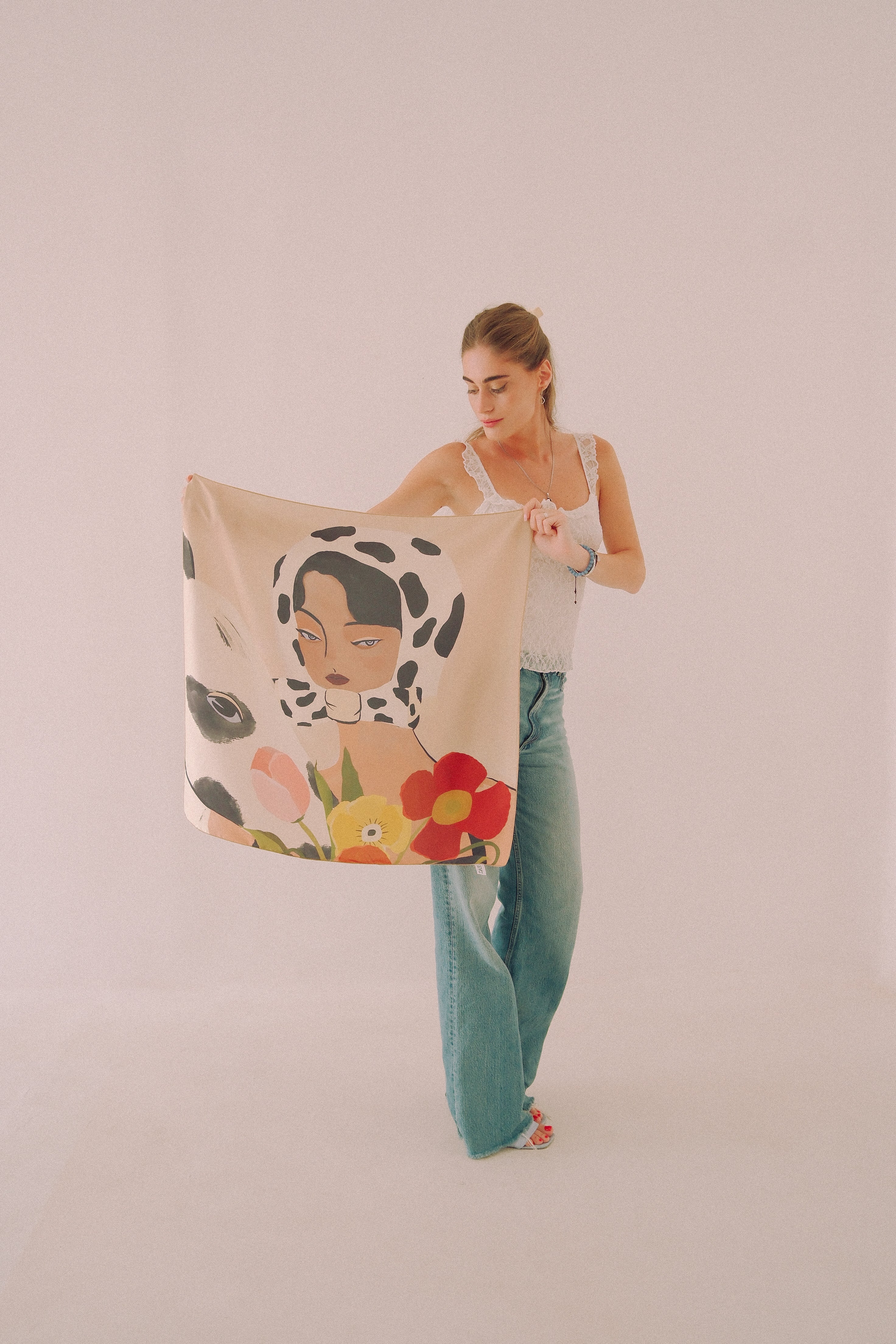 A woman holds up a large square scarf featuring a stylized illustration of a woman's face on a black-and-white spotted background, surrounded by colorful flowers, against a plain light pink studio backdrop.