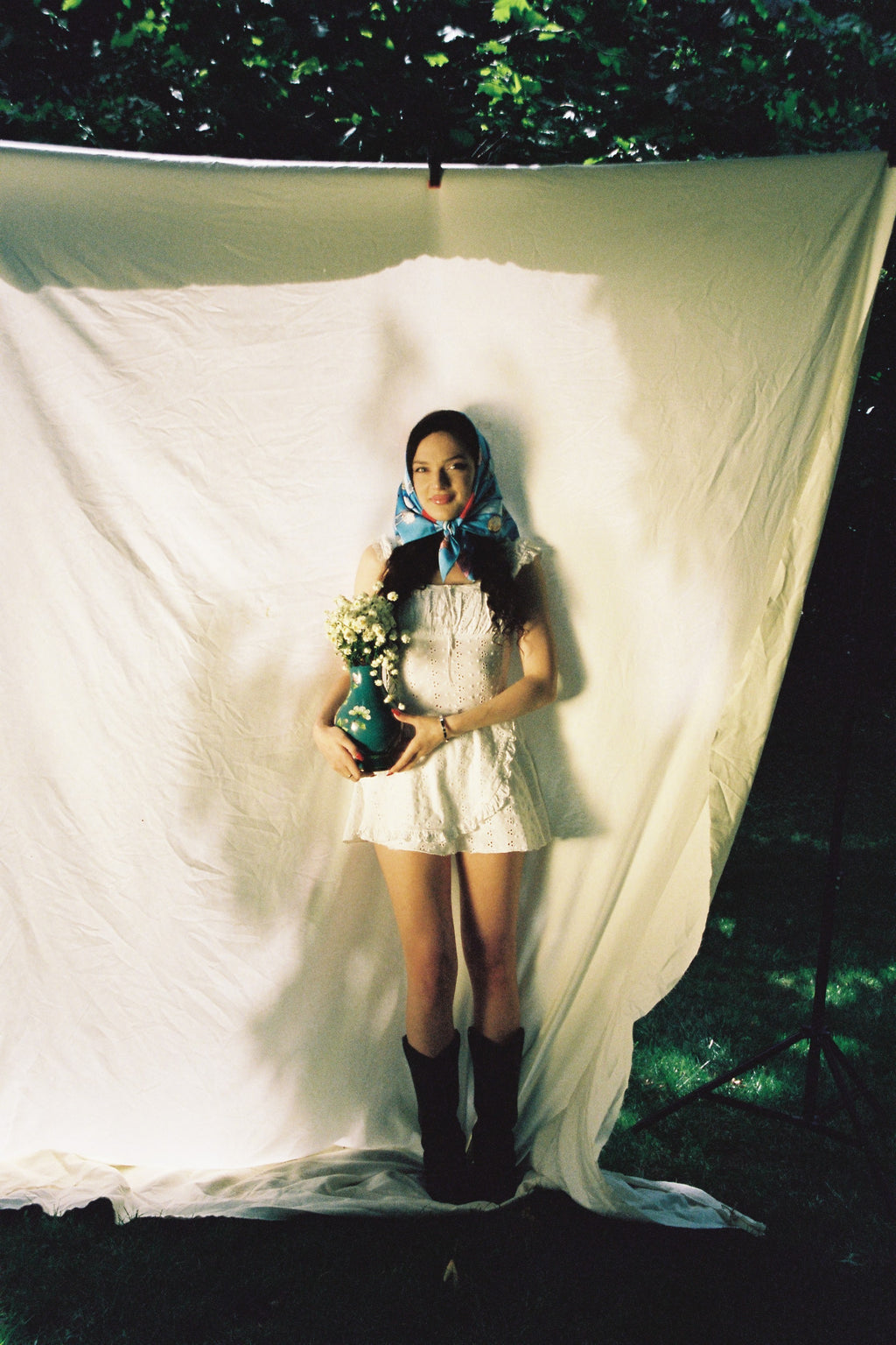 A woman lies on a large white sheet outdoors, wearing a white dress, blue top, and black boots, while holding a bouquet of white flowers, wearing scarf of DAE