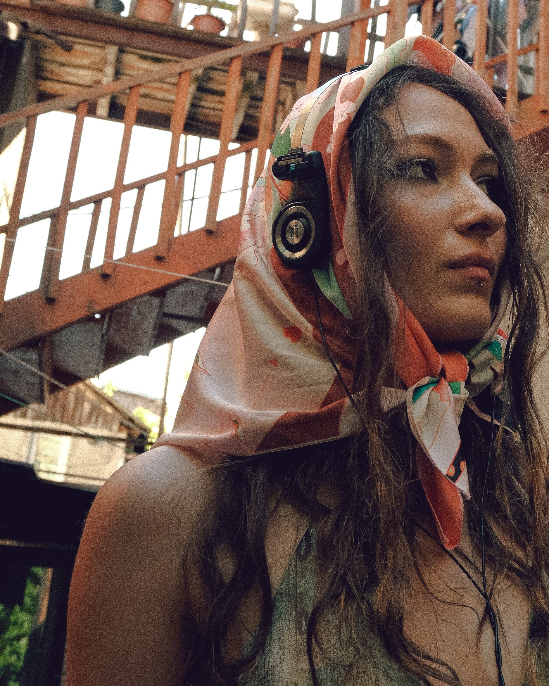 Woman wearing a colorful headscarf with a camera lens design, standing in an outdoor setting.