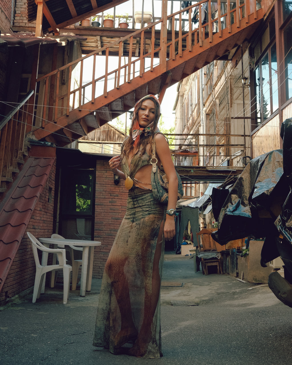 Woman in a long, patterned dress standing in an outdoor setting with a staircase and tables in the background, wearing scarf by DAE x Leah Han