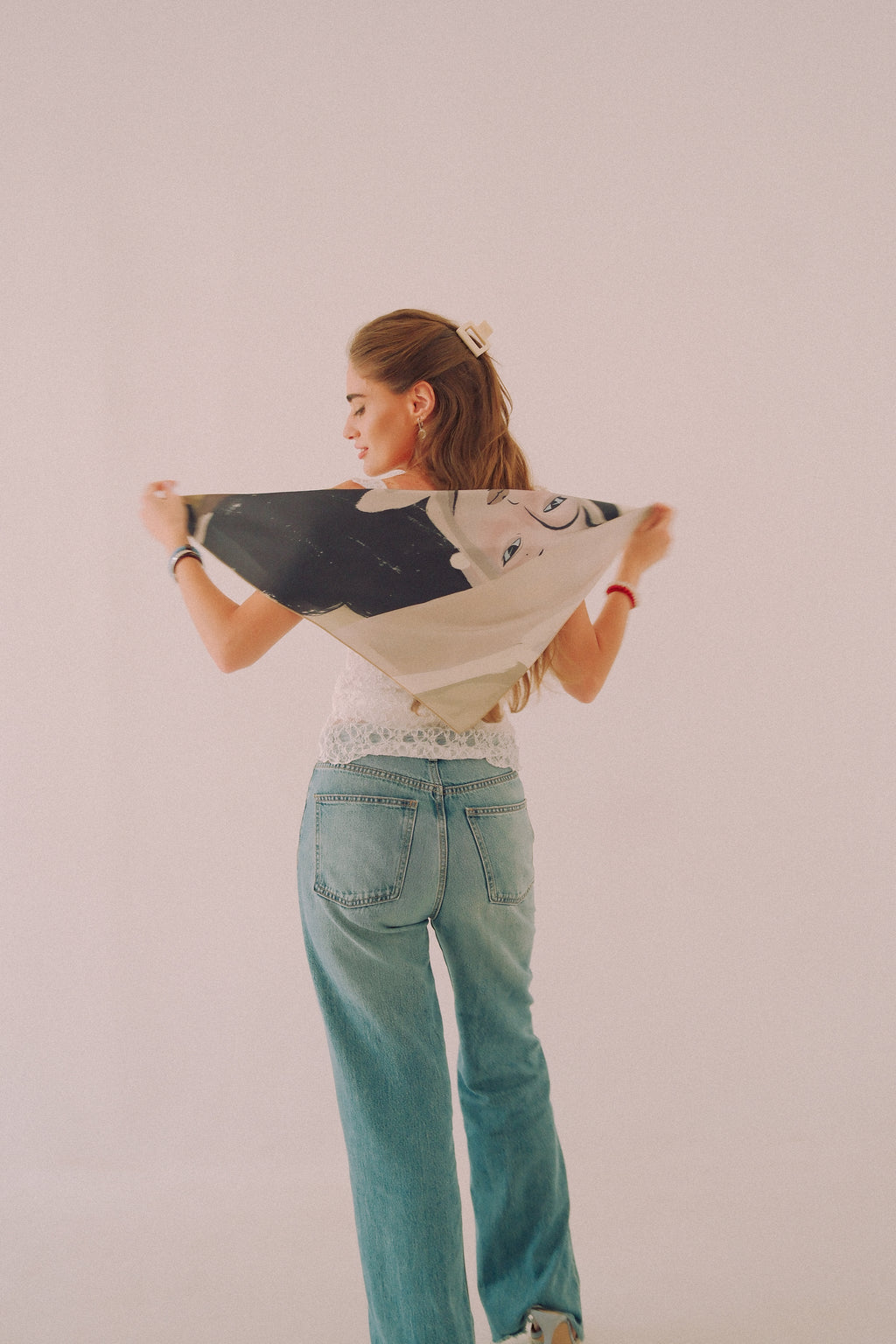 A woman stands with her back to the camera, holding up a black and white patterned scarf by DAE. She is wearing a white lace-trimmed top and light blue jeans, set against a plain, warm off-white studio background.