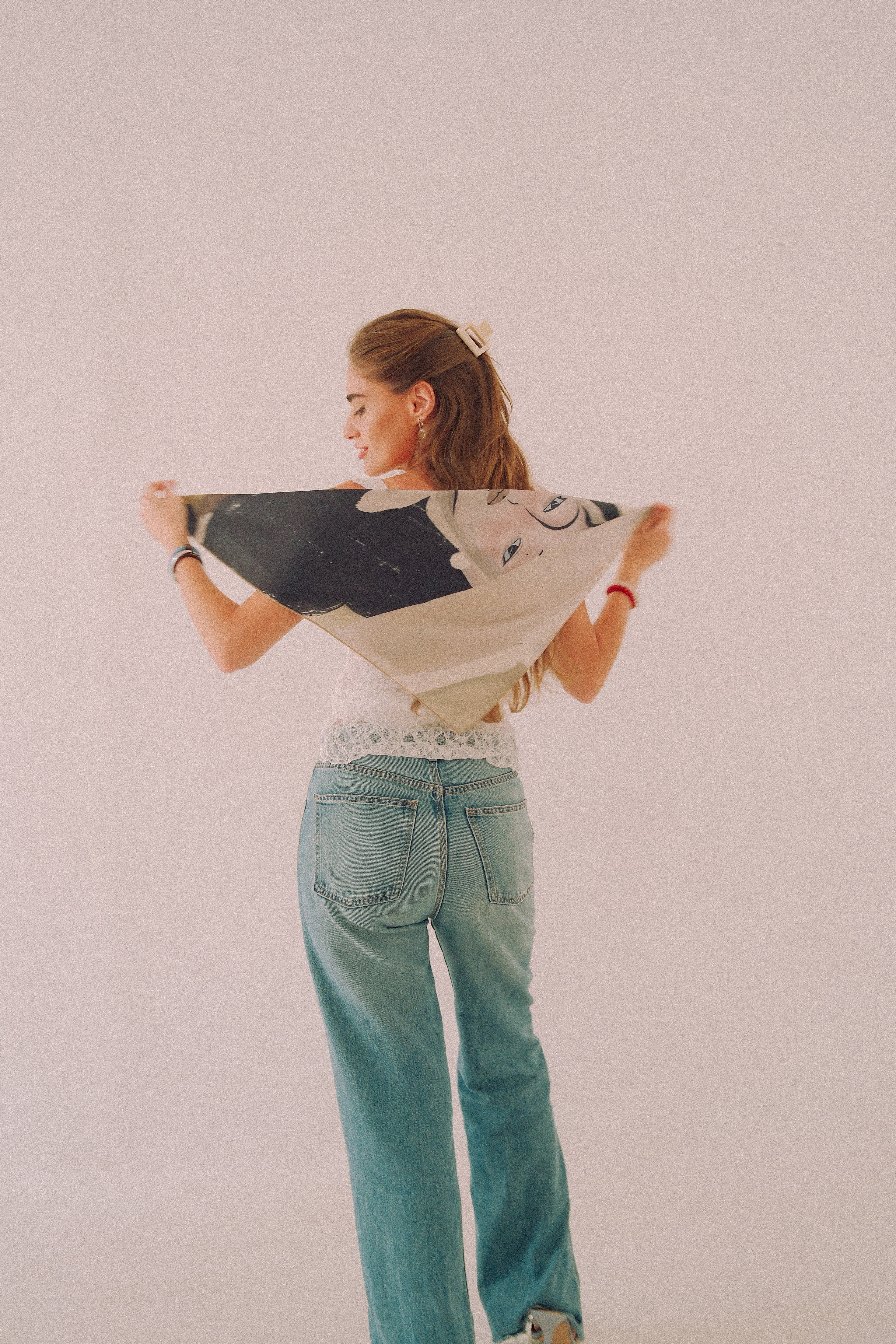 A woman stands with her back to the camera, holding up a black and white patterned scarf by DAE. She is wearing a white lace-trimmed top and light blue jeans, set against a plain, warm off-white studio background.