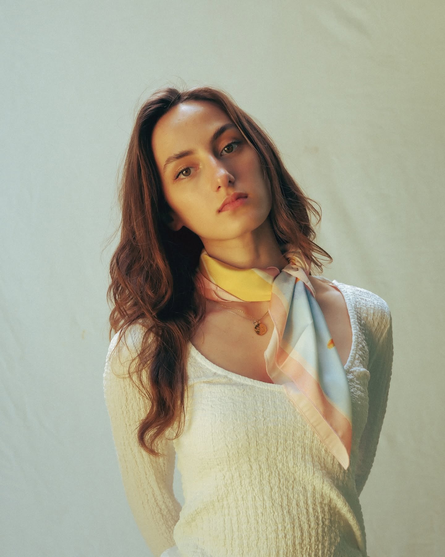 A woman with long wavy brown hair wearing a cream-colored textured knit top and a pastel striped scarf, posing against a light beige backdrop.