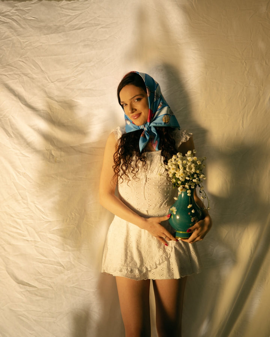 A woman wearing a white lace dress and a blue patterned headscarf stands against a draped cream fabric backdrop, holding a small green vase filled with white flowers.
