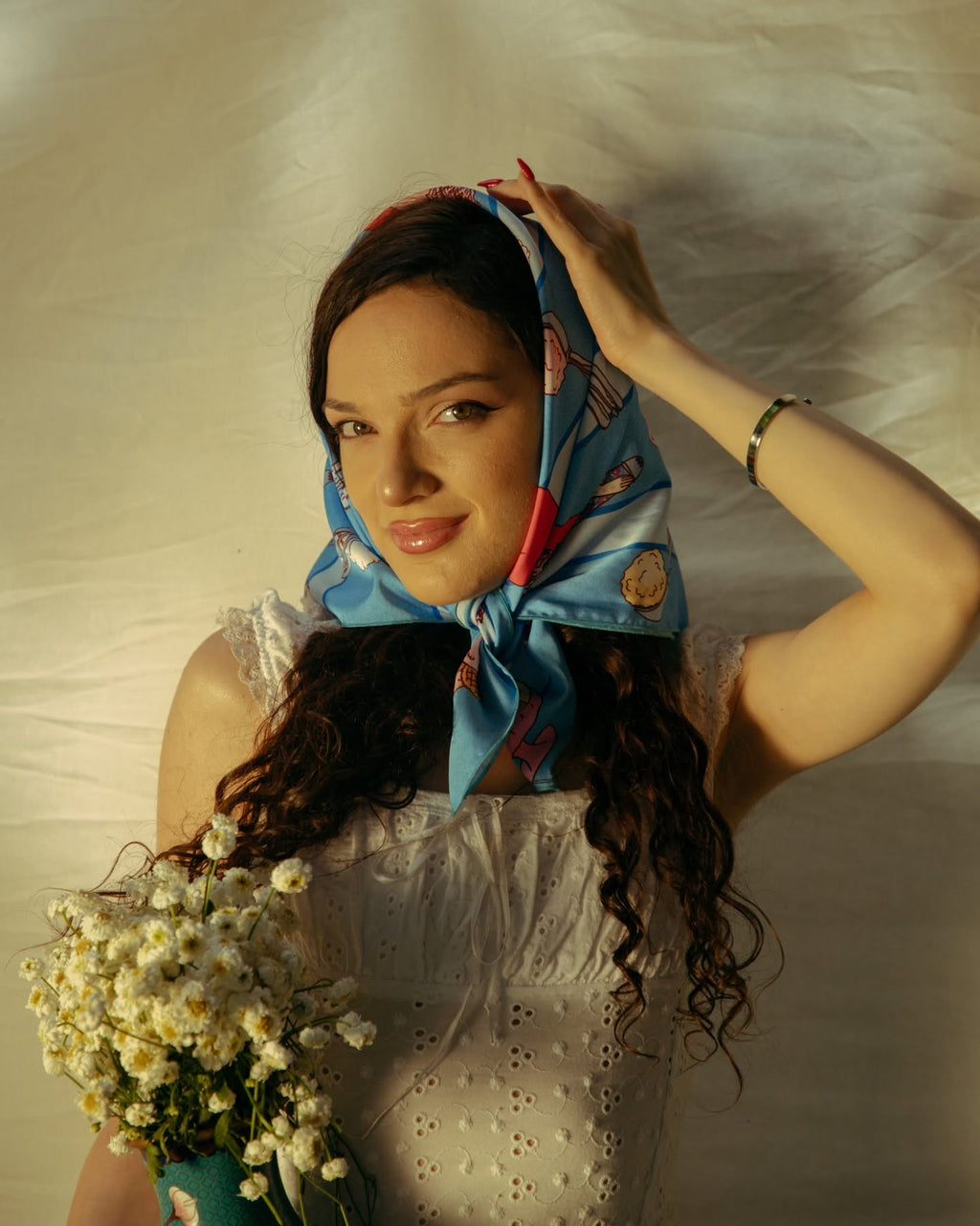 A woman wearing a light blue patterned silk scarf tied around her head, holding a bouquet of white daisies against a warm, draped cream fabric background.