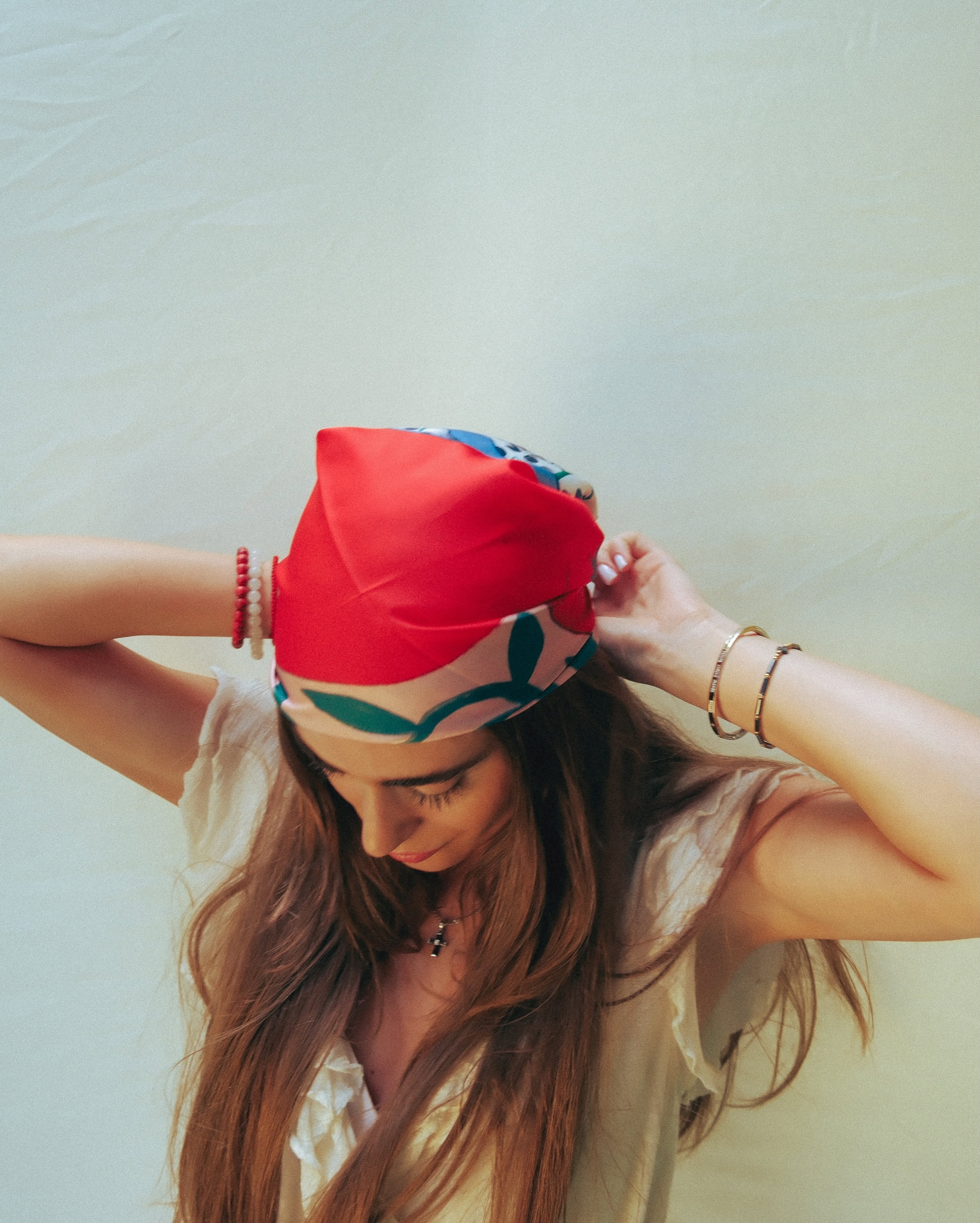 A woman with long brown hair wearing a red and multicolor patterned fabric headscarf, adjusting it with both hands against a plain light background.