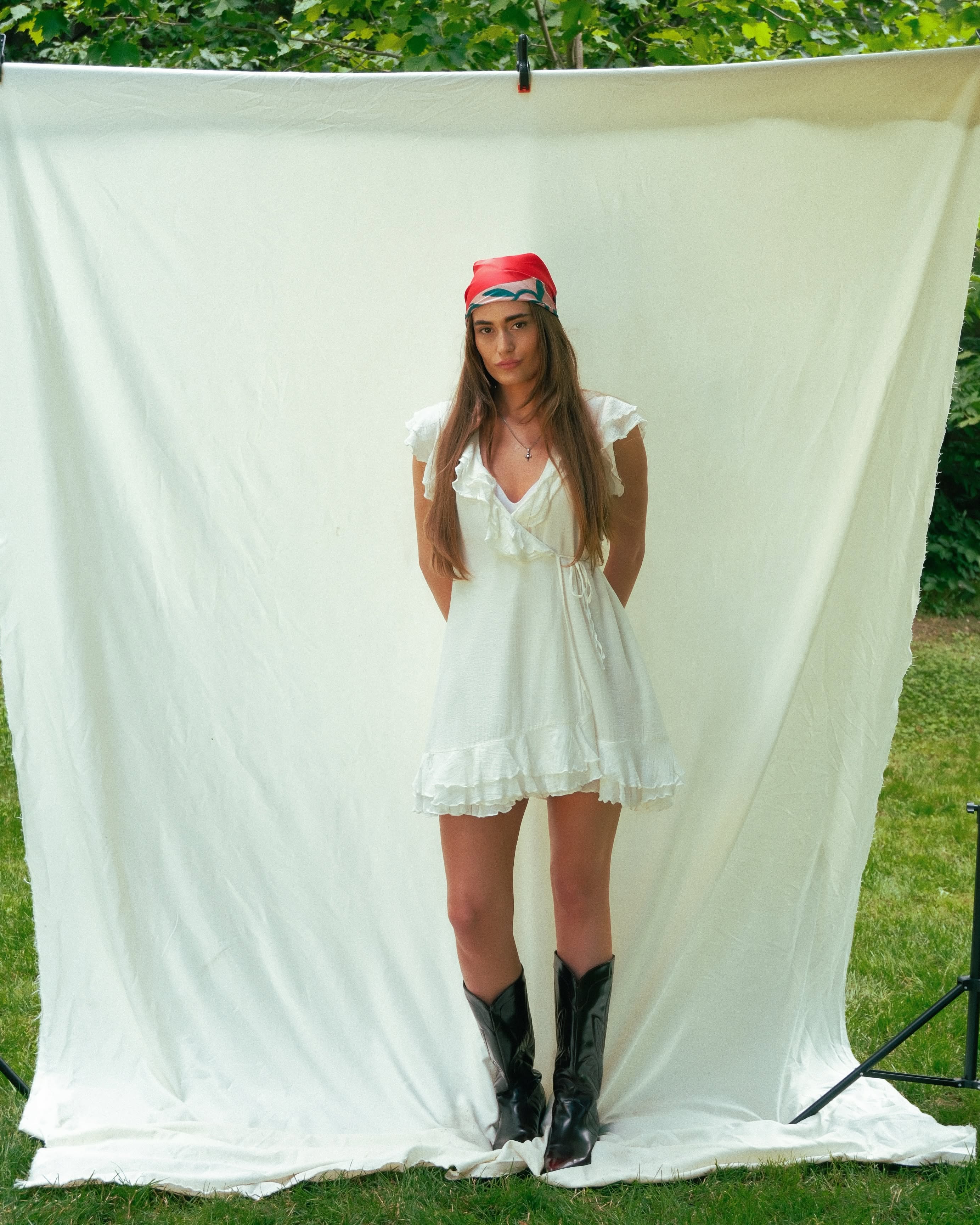 A woman stands outdoors in front of a large white fabric backdrop, wearing a white ruffled mini dress, a red scarf and black cowboy boots.