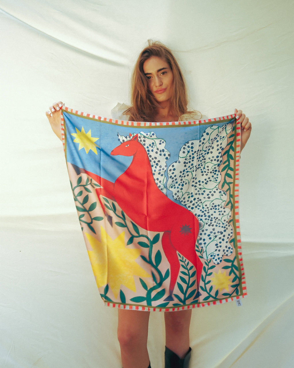 A woman holds up a square silk scarf featuring a vibrant, folk-art style illustration of a red horse, spotted deer, green leaves, and yellow suns on a blue and green background.