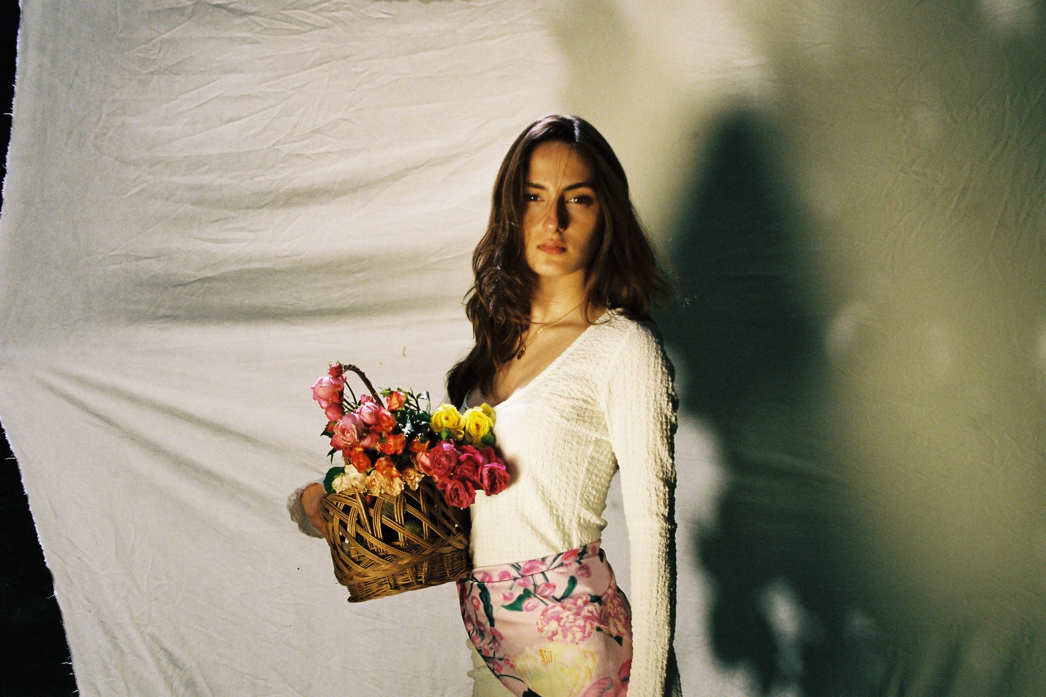 A woman stands against a draped white fabric backdrop holding a wicker basket filled with colorful flowers. She is wearing a white long-sleeve top and a pink floral skirt.