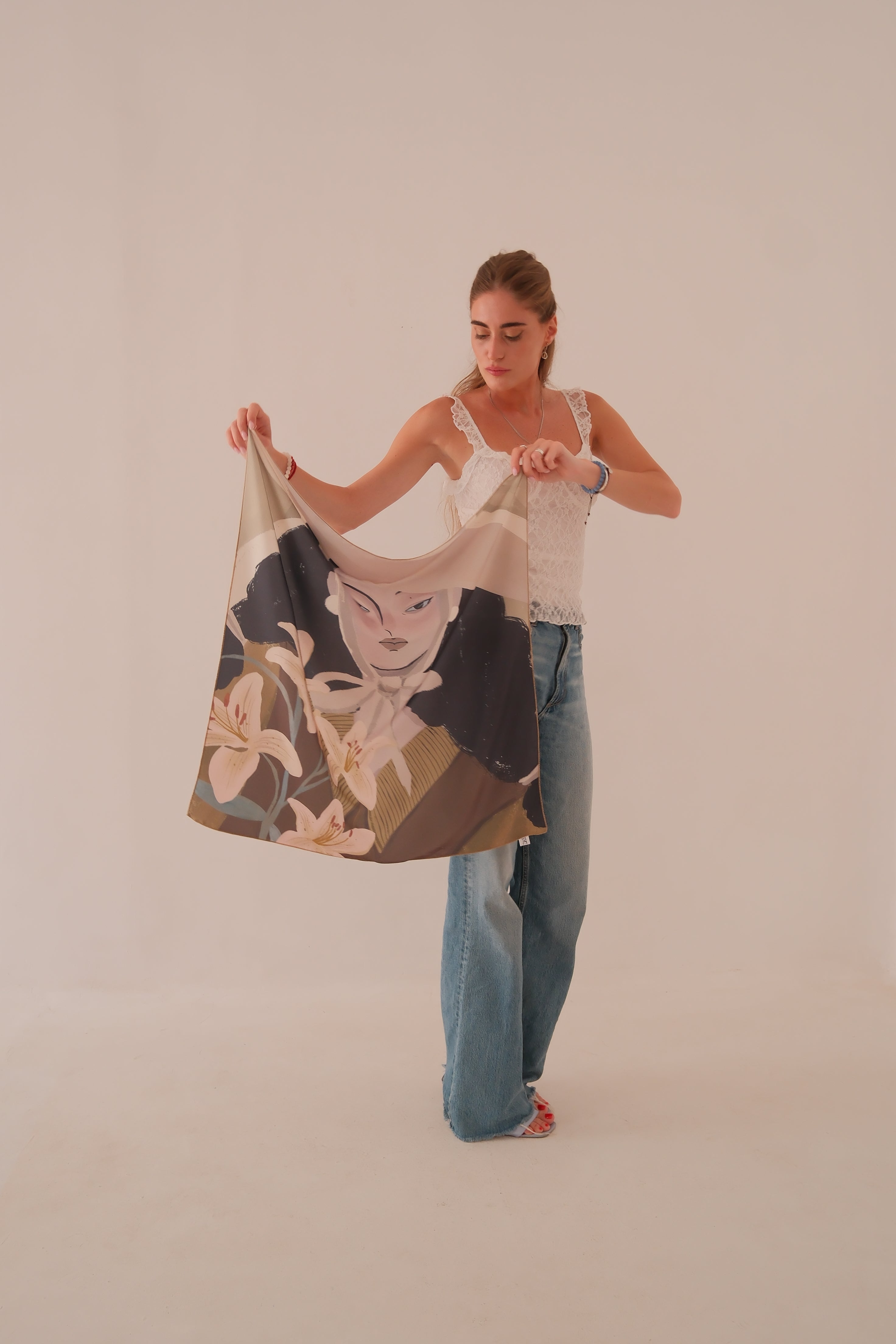 A woman stands against a plain beige studio background holding up a large scarf by DAE. The scarf features a printed illustration of a woman's face with floral elements in beige, brown, black, and cream tones.
