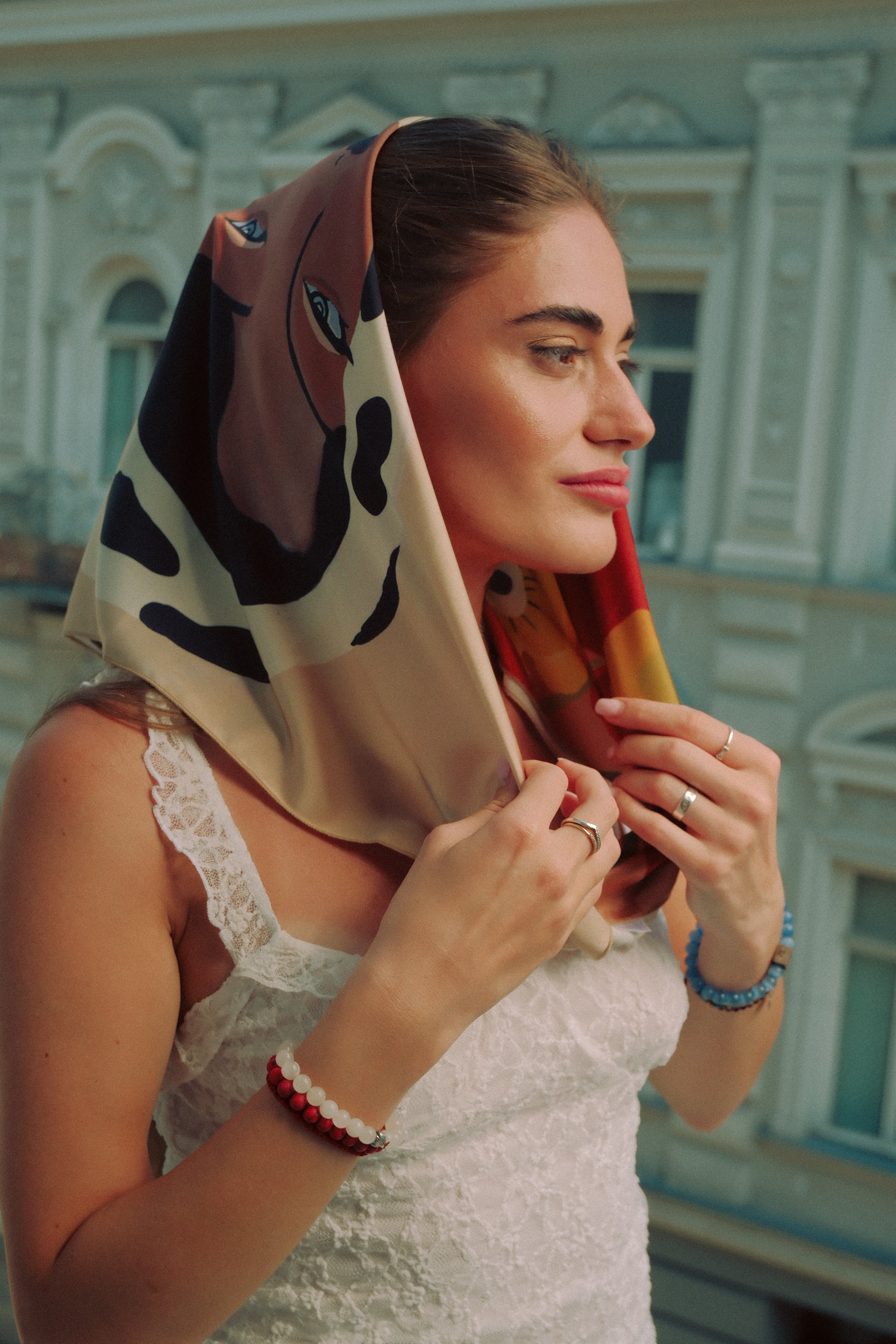 A woman wearing a beige silk scarf with a black and brown graphic print and a red patterned edge, styled over her head as a headscarf. She is wearing a white lace-trimmed top, a red and white beaded bracelet, and a blue beaded bracelet, with a blurred building facade in the background.